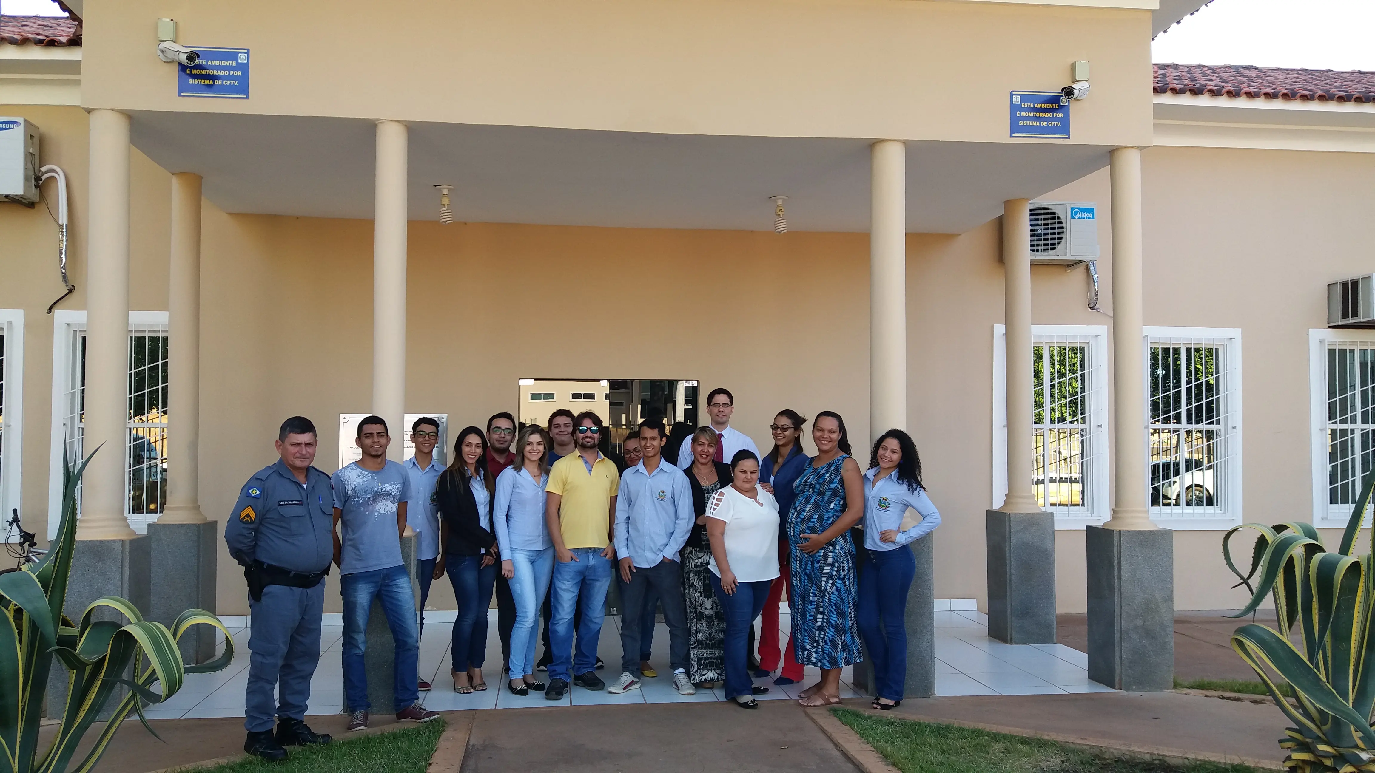 Equipe da Comarca de Campinápolis posa sorridente na entrada do prédio. O grupo, composto por servidores e magistrados em trajes casuais, demonstra integração da equipe em frente à fachada moderna com colunas e sistema de monitoramento, evidenciando ambiente acolhedor de trabalho.