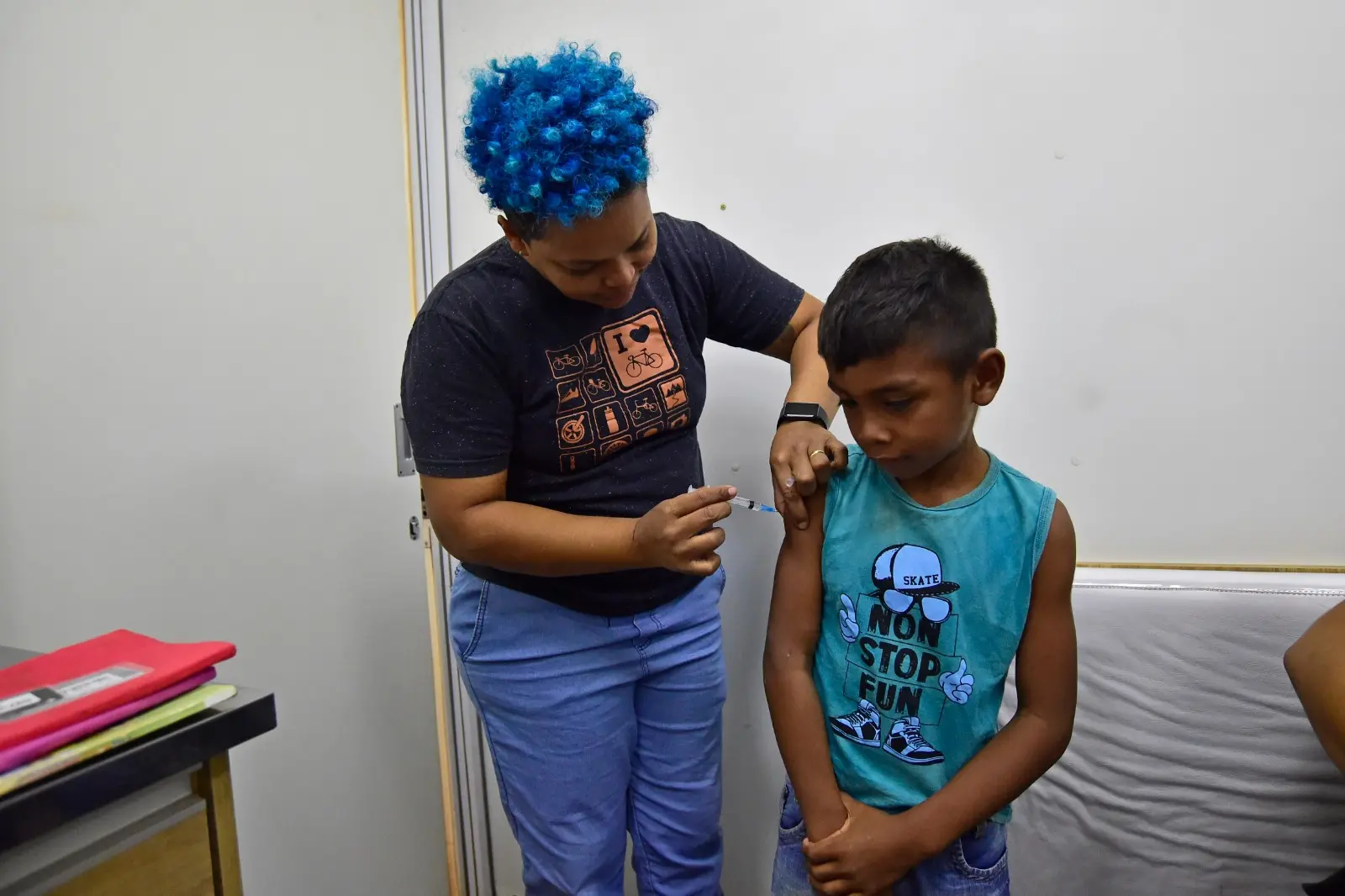 Menino recebe vacina no braço dentro de unidade móvel. Técnica de cabelo azul aplica a dose com atenção. O garoto observa o procedimento com expressão séria, vestindo camiseta azul.