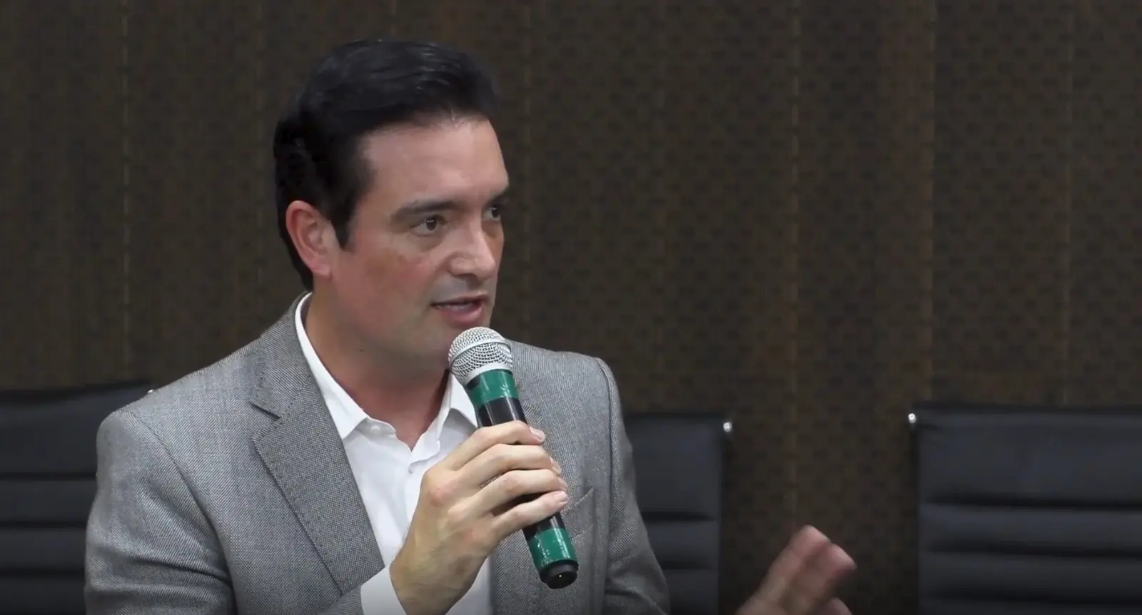 Leonardo Bertolin  é um homem de cabelos escuros e penteados, vestindo um blazer cinza e camisa branca aberta, fala em um microfone verde e cinza. Ele gesticula com a mão direita, em um ambiente interno com paredes marrons.