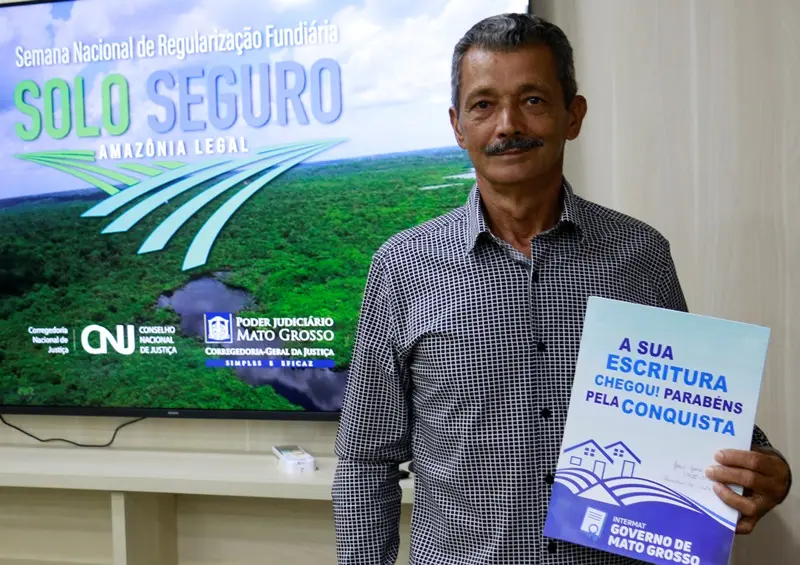 a foto retrata o morador em pé, ele segura uma pasta de papel que contém o título de propriedade.