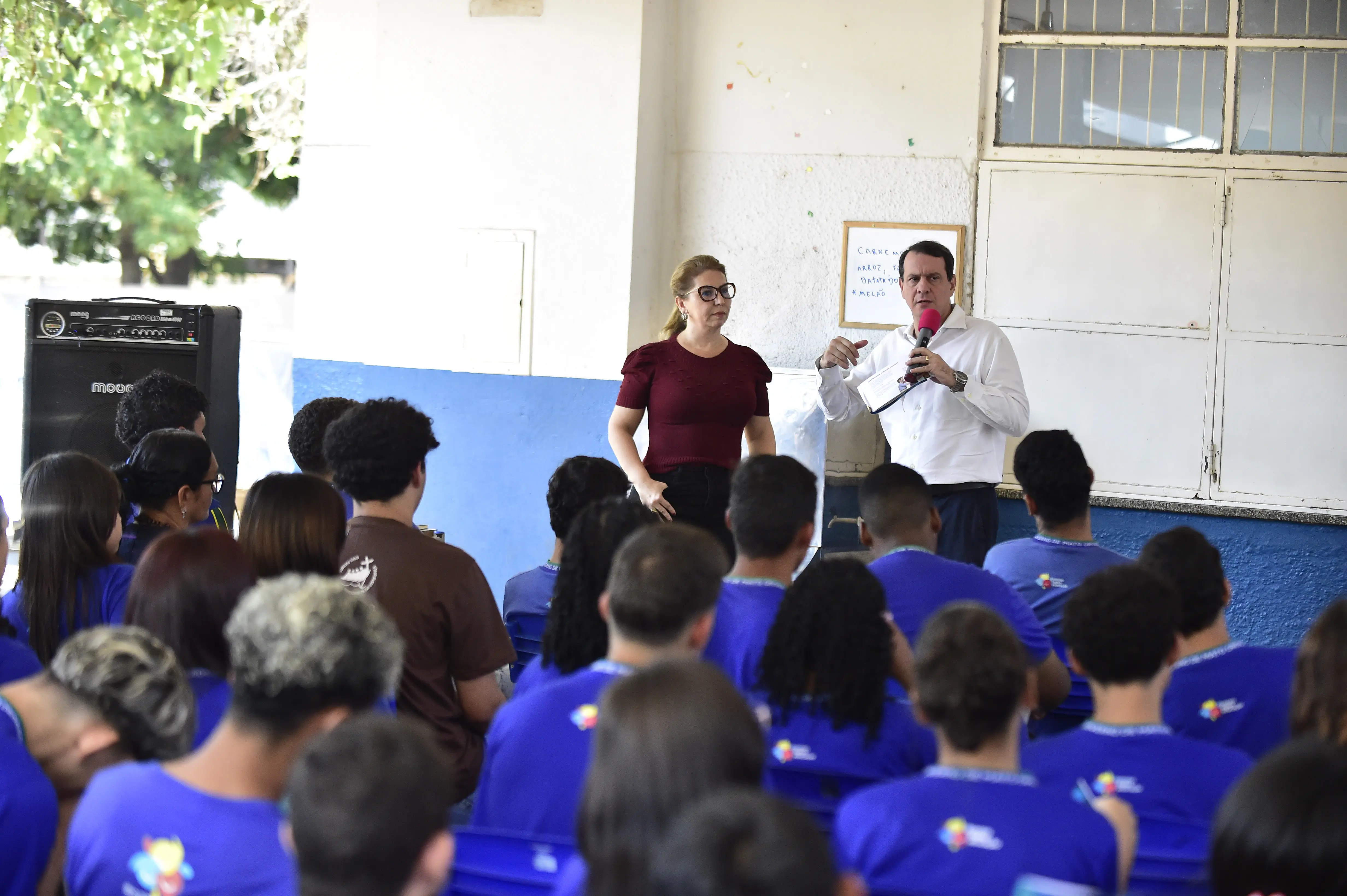 Homem com microfone e mulher em pé falam a estudantes de uniforme azul em auditório escolar, durante atividade educativa do projeto Nosso Judiciário na Escola, promovendo diálogo e orientação aos jovens.