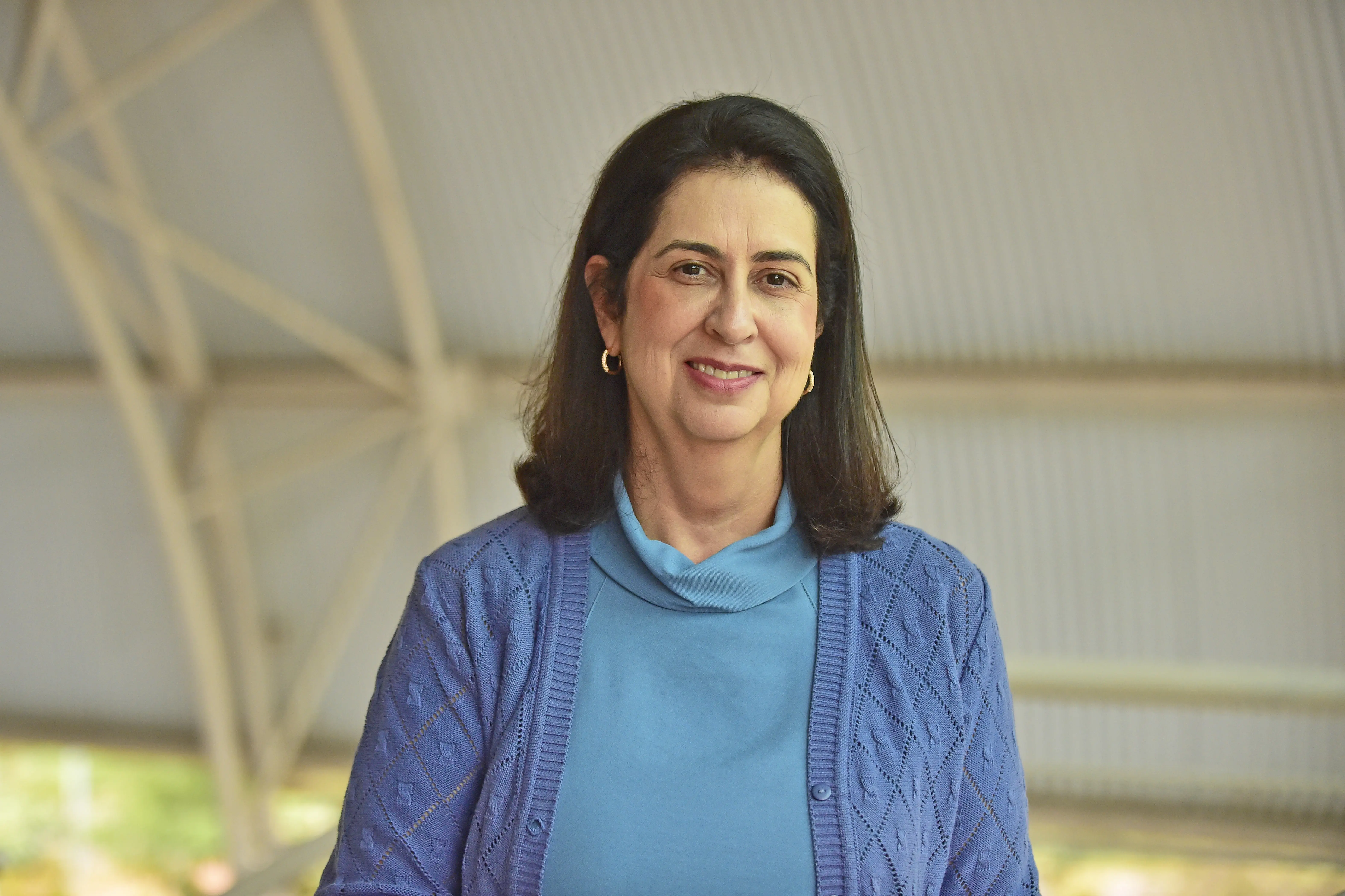 Uma mulher de pele clara, cabelo escuro na altura dos ombros, veste uma blusa azul com um casaco de tricô azul claro e brincos de argola dourados, sorrindo gentilmente