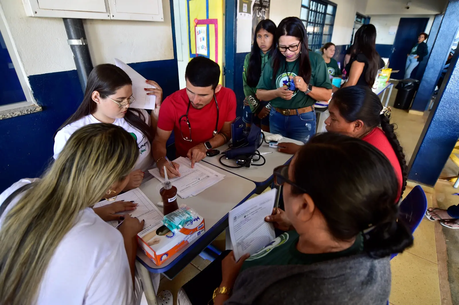 Profissionais de saúde e comunidade indígena preenchem fichas e realizam atendimentos médicos em ambiente escolar, com participação ativa de mulheres.