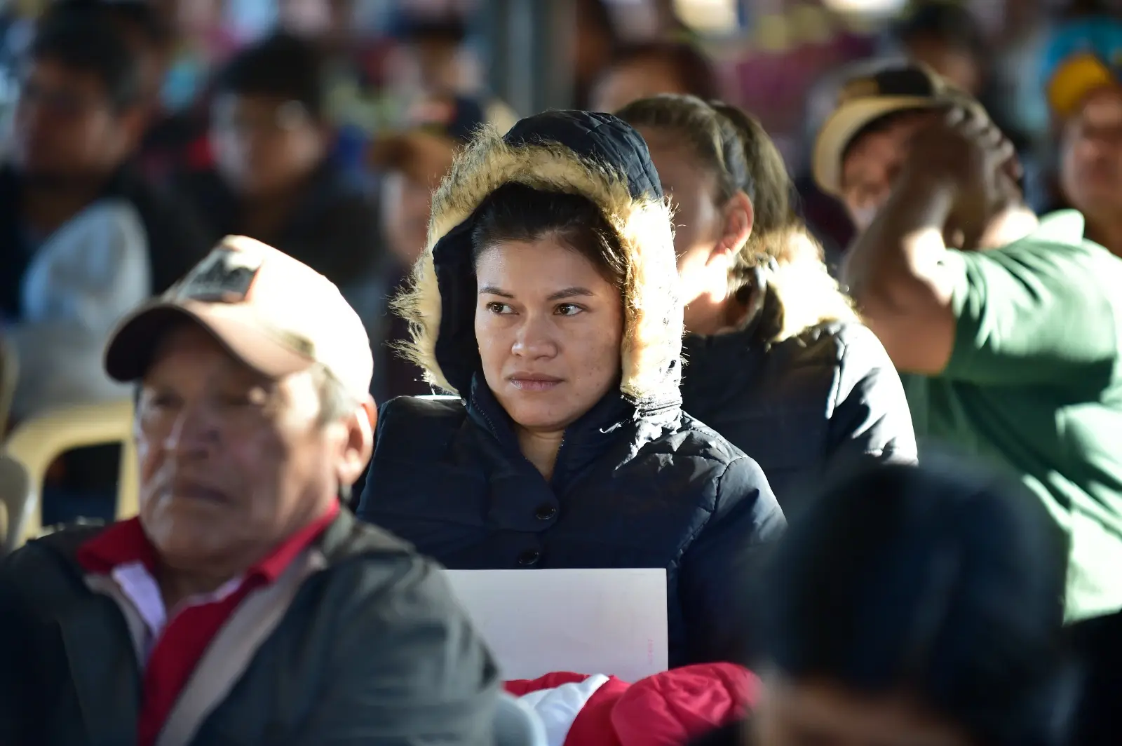 Mulher com jaqueta de frio observa atenta uma atividade. Ela está sentada entre outras pessoas, em local coberto, durante ação comunitária da Justiça Sem Fronteiras.