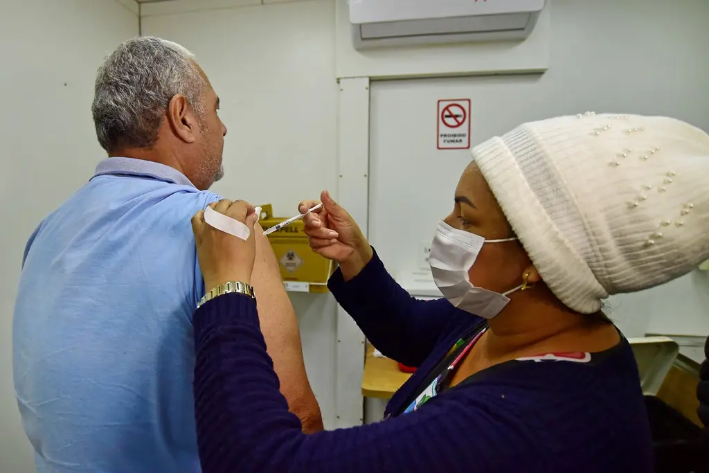 Homem de costas, usando camiseta polo azul celeste é vacinado no braço por uma mulher que usa máscara.