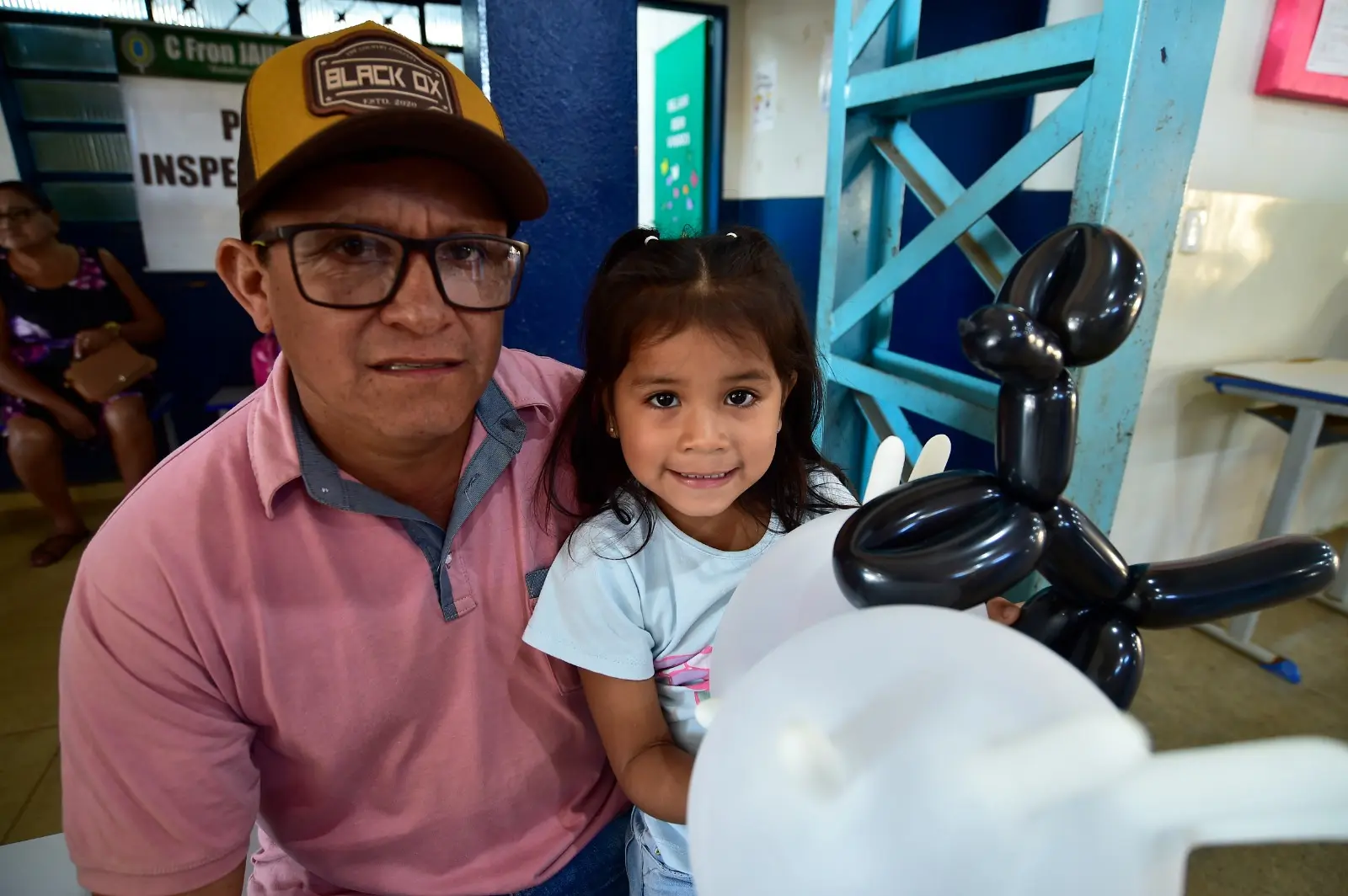 Homem com boné segura criança sorridente no colo. Eles estão sentados em sala de espera do posto de saúde. A menina segura uma escultura de balão em forma de cachorro preto e branco.