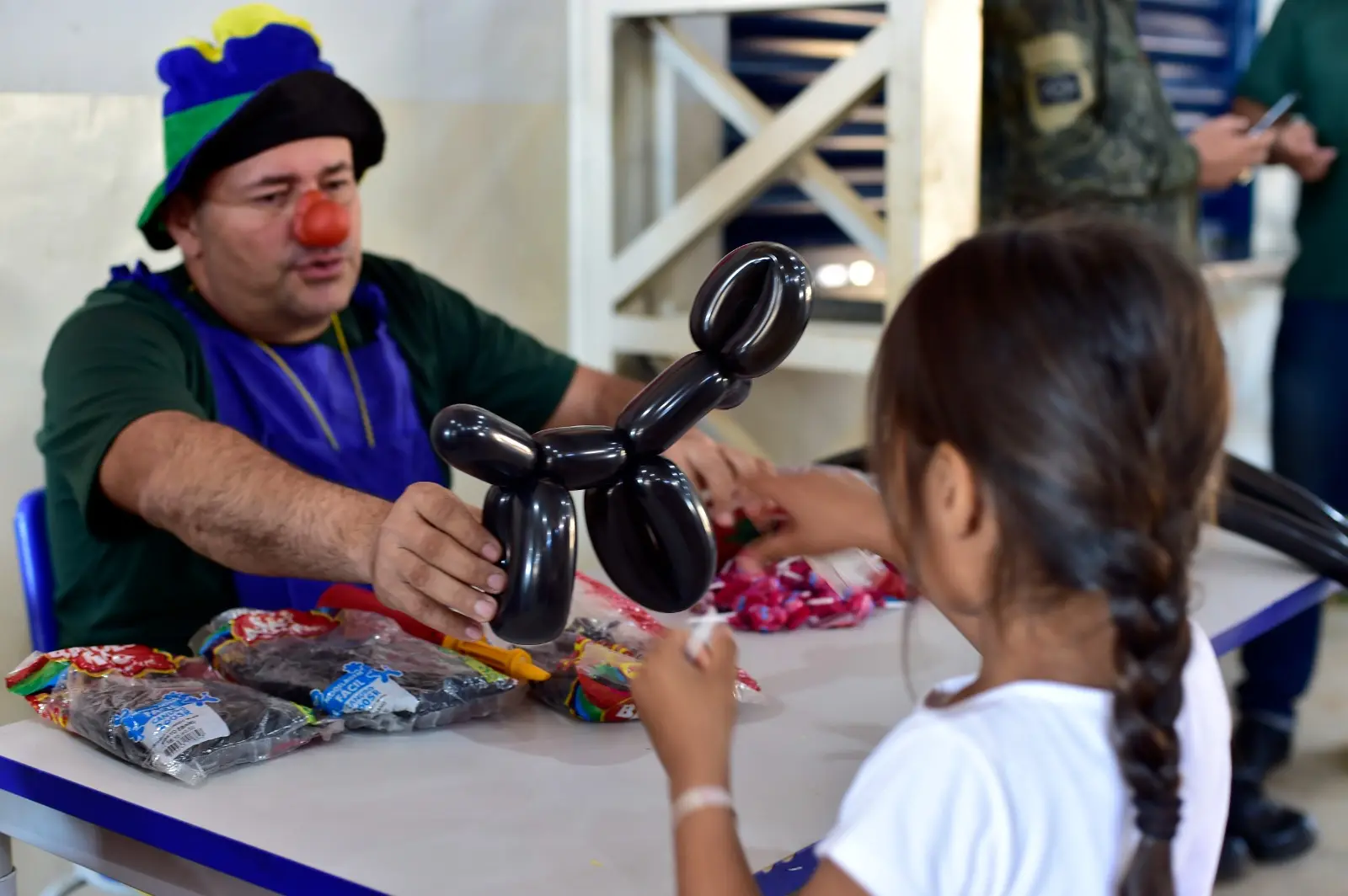 Homem fantasiado de palhaço entrega escultura de balão para uma menina. Em cima da mesa, há vários balões ainda embalados e um pacote de doces.