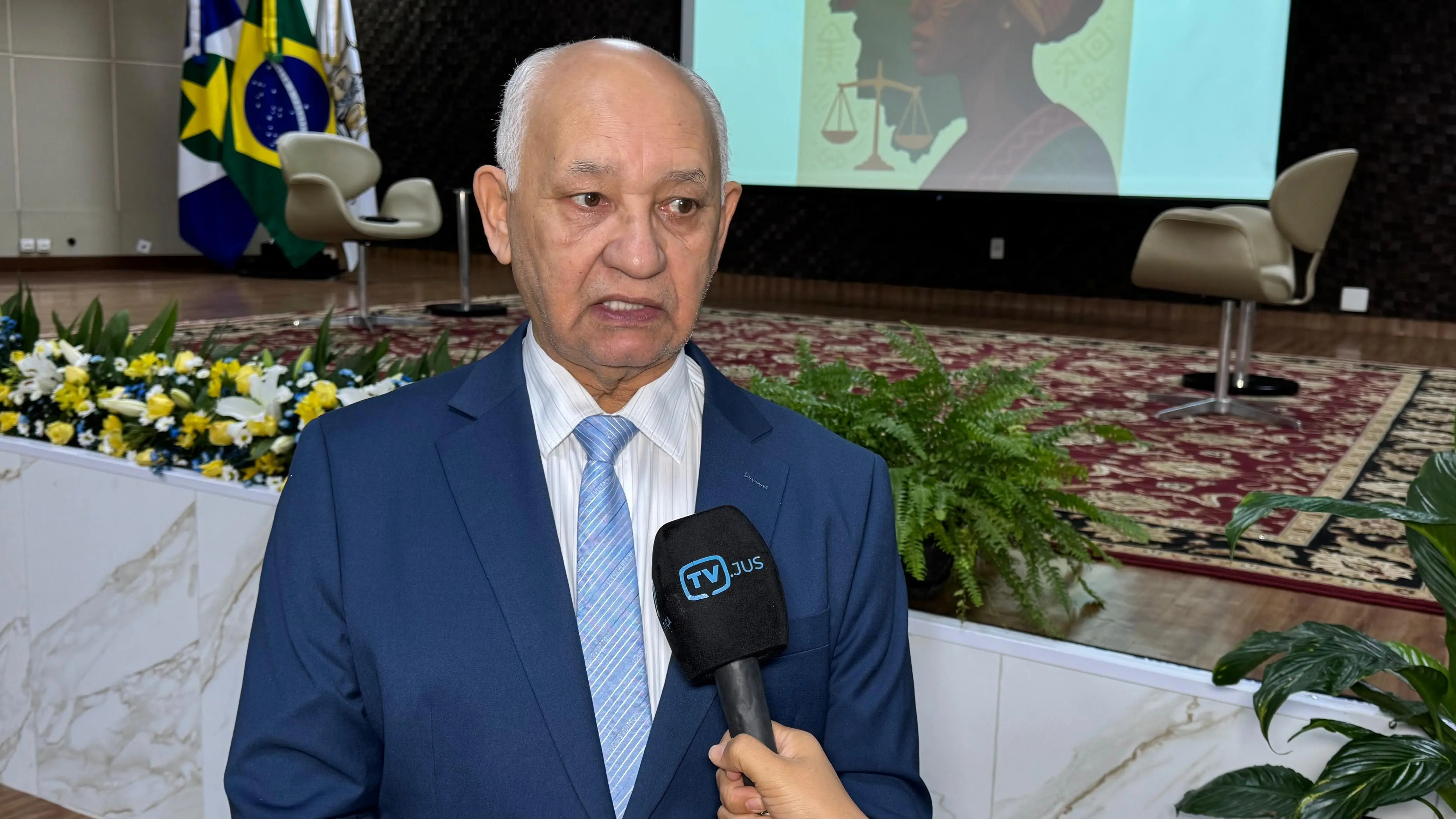 Desembargador Juvenal Pereira da Silva concede entrevista à TV.Jus. Ele é um senhor de pele parda, calvo, de cabelos brancos, olhos castanhos, usando camisa branca listrada, gravata azul clara listrada e terno azul. 