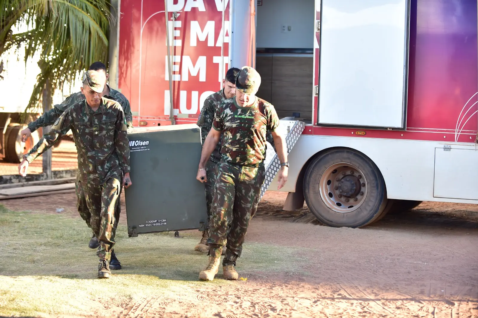 Foto que mostra quatro militares do Exército carregando uma enorme caixa de metal, que acabaram de descer da carreta, que aparece ao fundo.