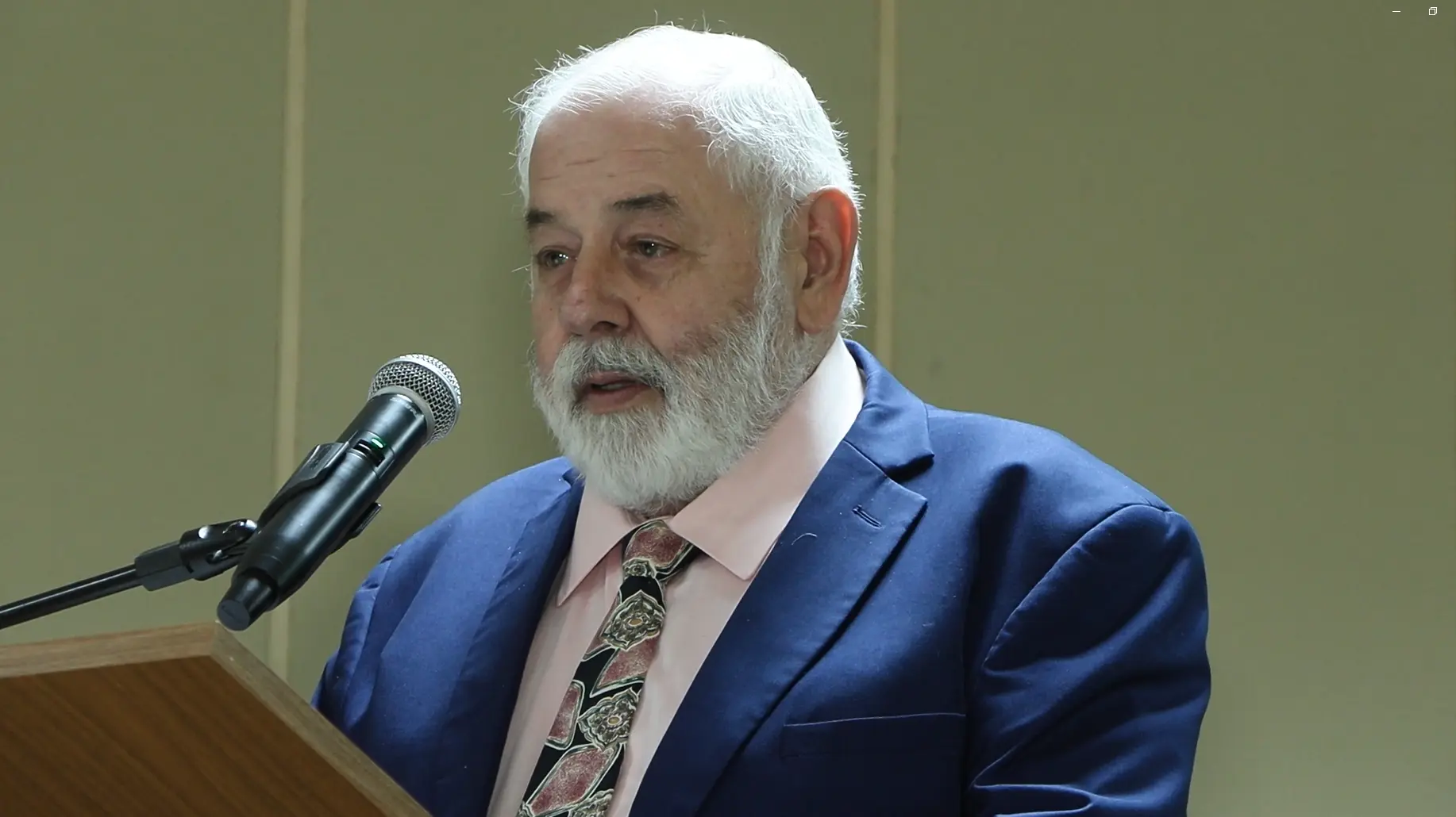 Zuquim – Presidente do TJMT, desembargador José Zuquim, fala ao microfone. Ele é um senhor branco de cabelos e barba brancos, olhos claros, usando camisa rosa clara e terno azul. 