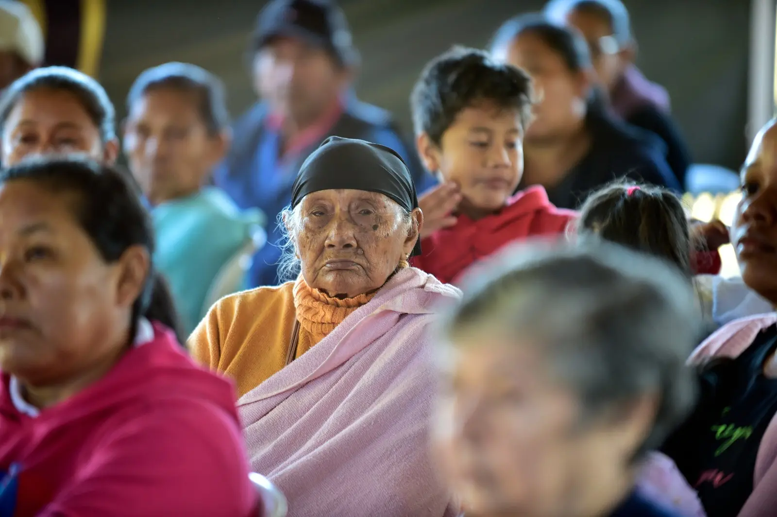 Idosa com lenço preto e manta rosa observa a atividade ao redor, cercada por outras pessoas. Ao fundo, há adultos e crianças acompanhando o evento.