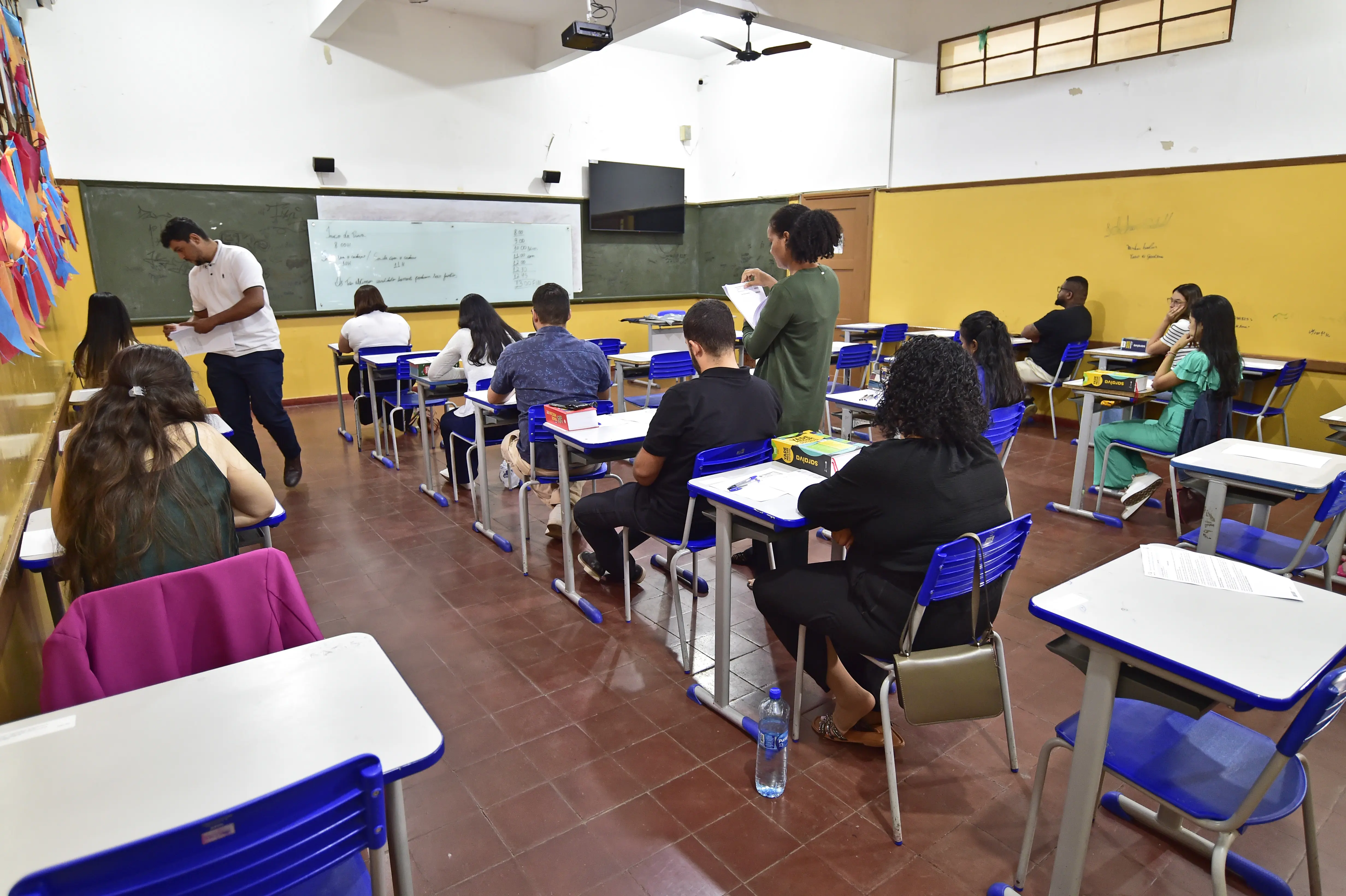 Foto que mostra a sala de aula com inscritos do processo seletivo sentados de costas para a foto, enquanto um homem e uma mulher distribuem as provas. 