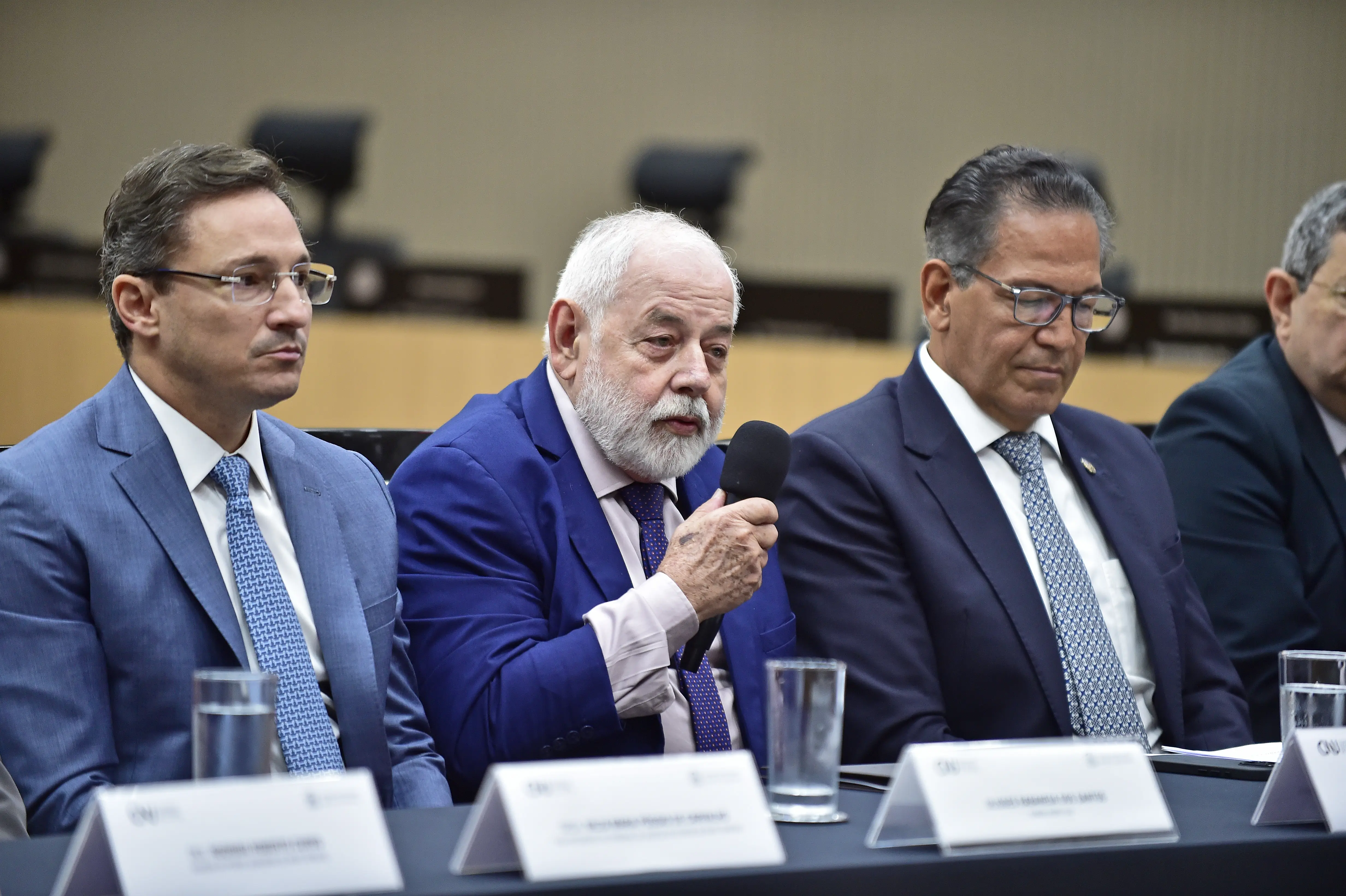 Presidente do TJMT, desembargador José Zuquim Nogueira, fala ao microfone, sentado entre o conselheiro do CNJ, Ulisses Rabaneda, e o ministro corregedor Mauro Campbell. Zuquim é um senhor branco, de cabelos e barba brancos, olhos claros, usando terno e gravata azul marinho.