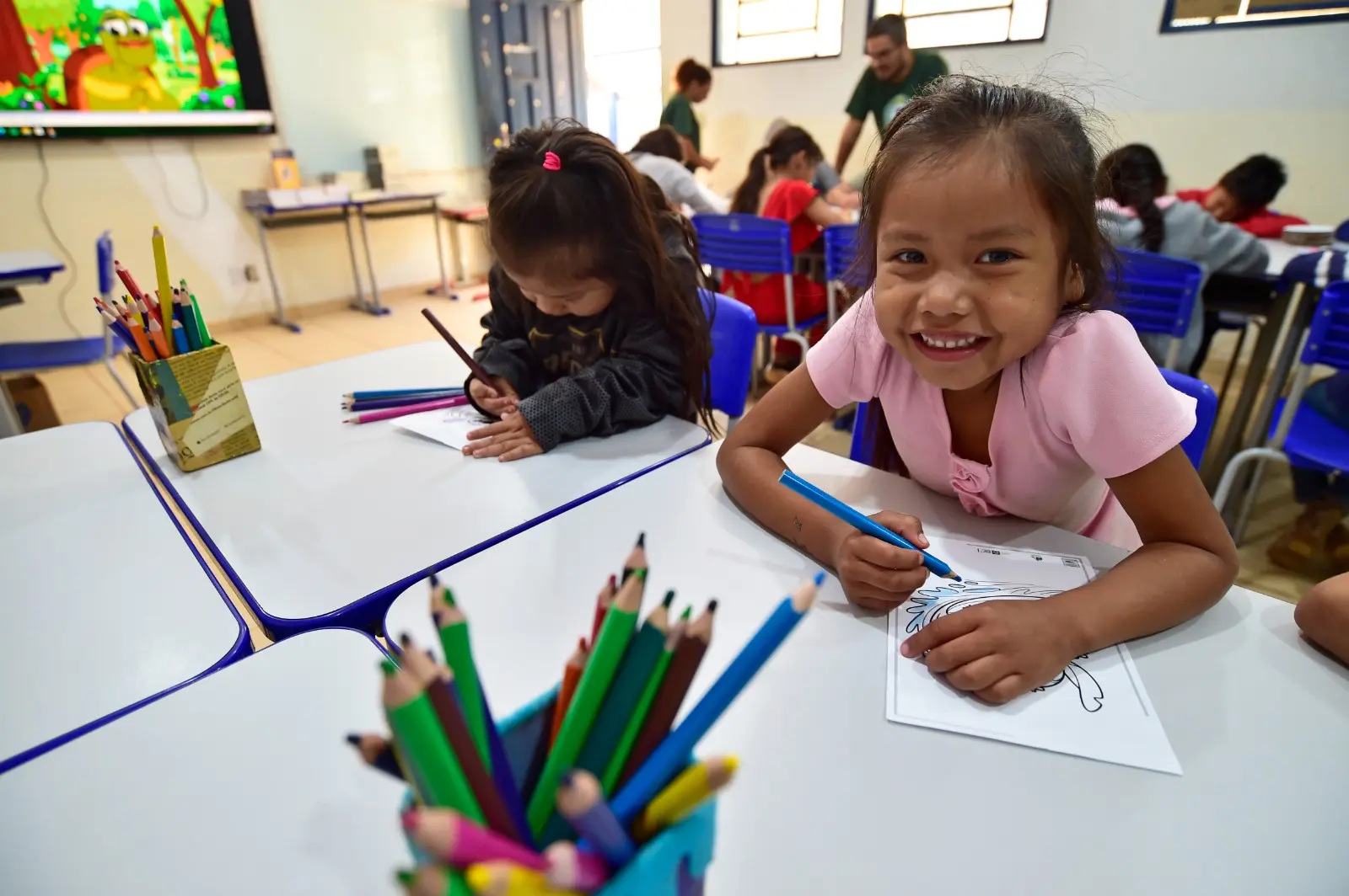 Criança sorri enquanto colore desenho com lápis azul em sala de aula. Ao lado, outra menina desenha concentrada. Há mais crianças ao fundo.