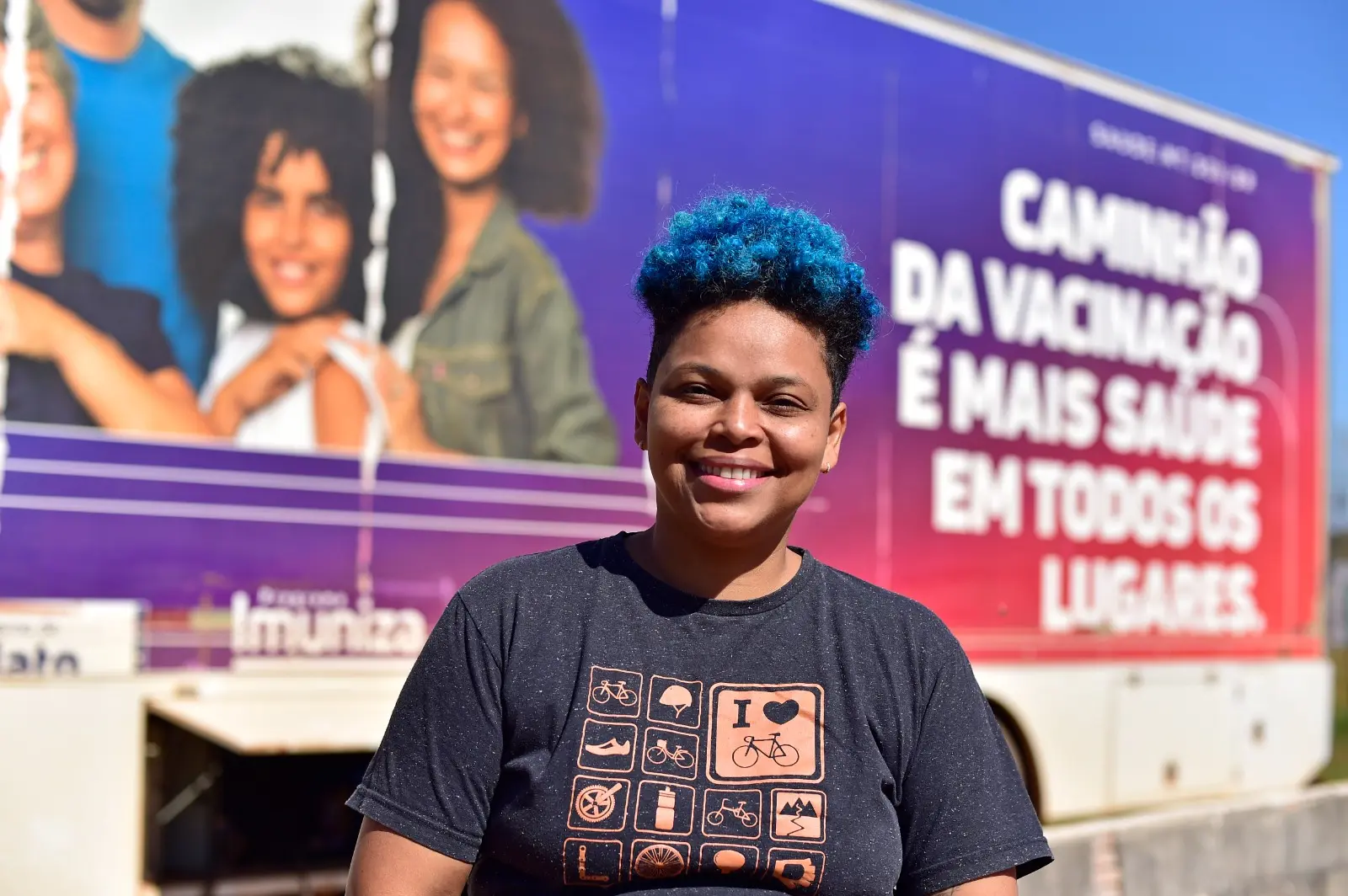 Profissional de saúde com cabelo azul sorri para a câmera. Ao fundo, painel do caminhão da vacinação traz mensagem sobre a importância da imunização em todos os lugares.