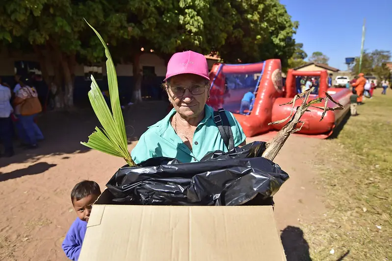 Foto que mostra uma idosa segurando uma caixa de papelão com mudas de plantas. Ao lado dela, há um menino de cerca de 3 anos. Eles estão no pátio de uma escola.