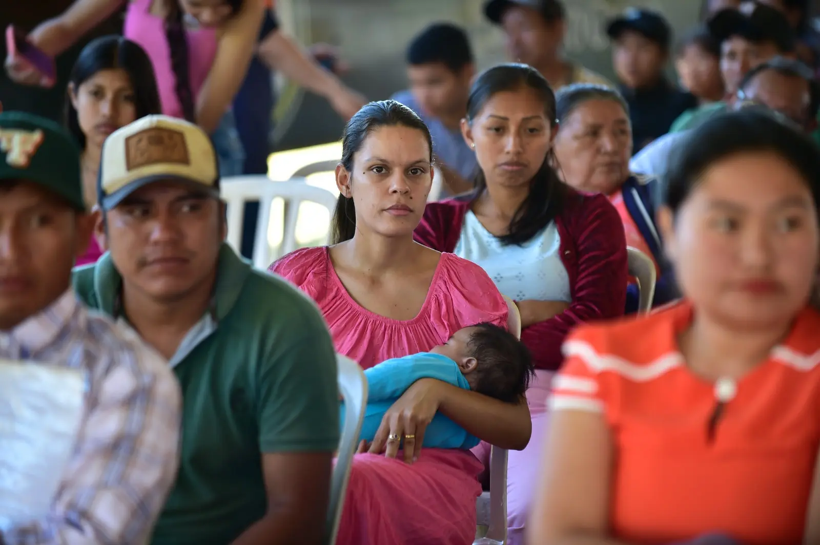 Mulher em destaque amamenta bebê no colo durante evento. Ela está sentada entre outras pessoas da comunidade, com expressões sérias, todas voltadas à frente, acompanhando atentamente