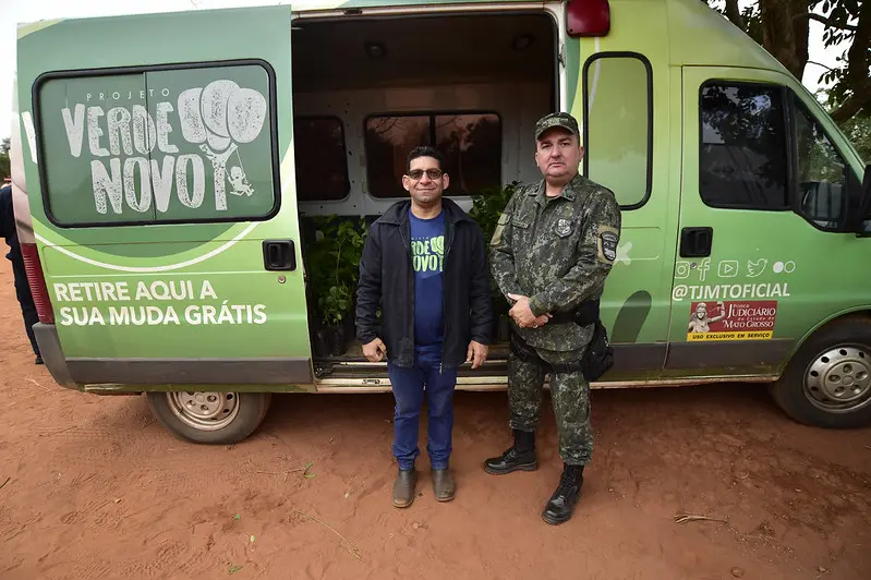 Foto que mostra o servidor do Verde Novo e o policial ambiental posando para a foto e sorrindo, em pé, em frente à van do programa Verde Novo. A porta da van está aberta e, dentro dela, há várias mudas de plantas.