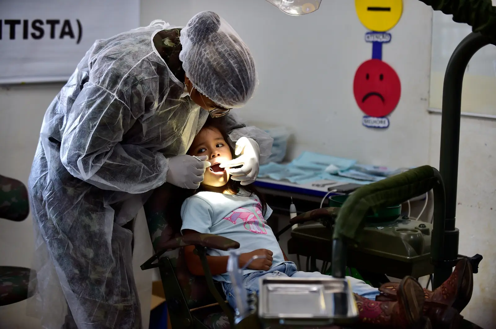 Dentista do Exército atende uma menina na cadeira odontológica. Ela está com uniforme do Exército e paramentos como touca, luva e capote.