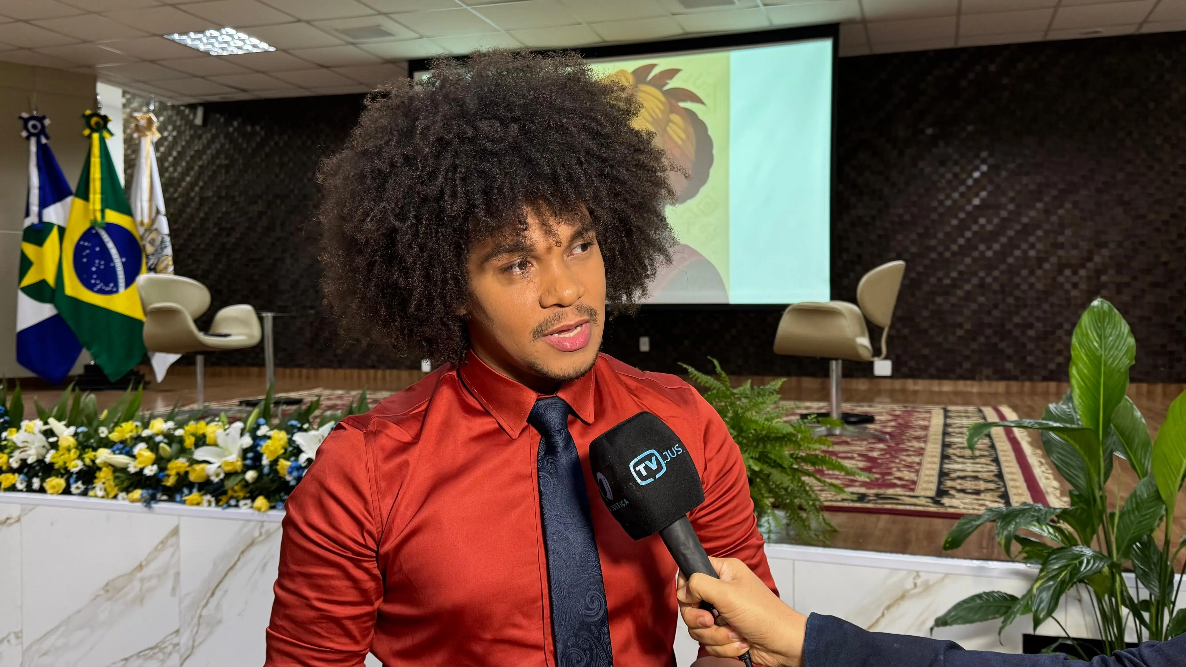Wagner Mônantha concede entrevista à TV.Jus. Ele é um jovem negro, de cabelo black power, olhos castanhos, usando camisa vermelha e gravata azul marinho.