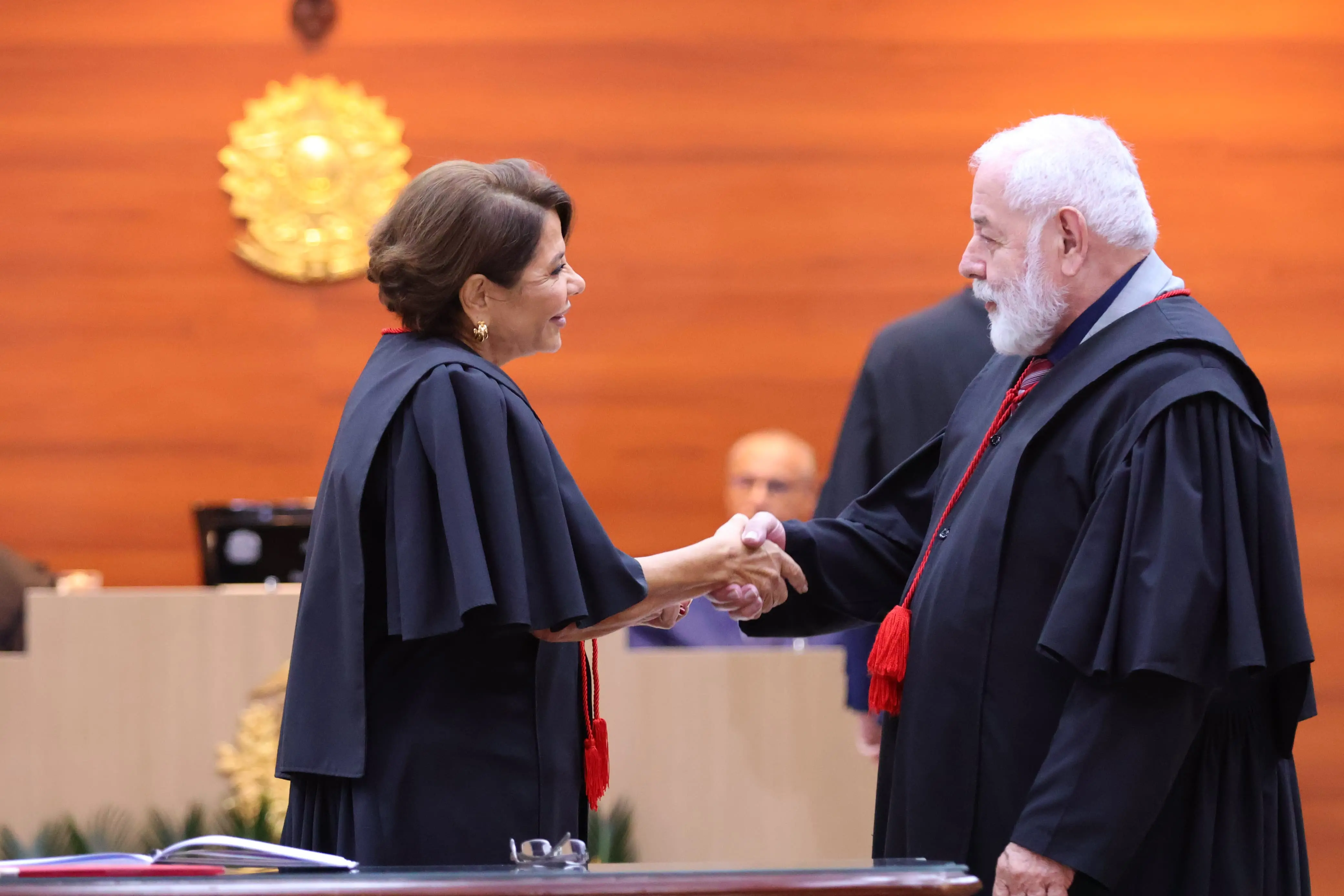 Mulher e homem, ambos com vestes talares pretas e detalhes em vermelho, apertam as mãos sorrindo em cerimônia oficial. Ao fundo, parede de madeira com símbolo da República e pessoas sentadas compõem o cenário solene.