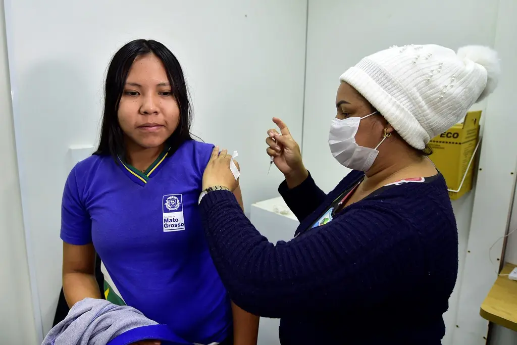 Adolescente menina com uniforme de escola estadual é vacinada por uma servidora que usa máscara, touca branca e blusa de frio azul marinho.