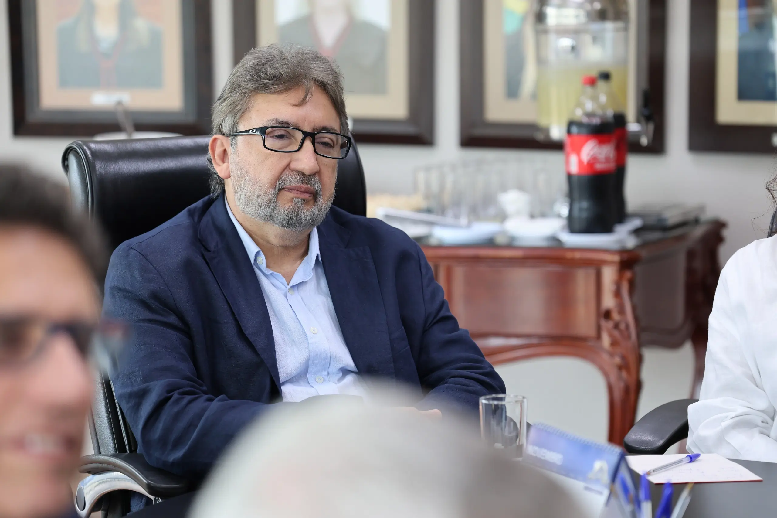Foto do des. Márcio Vidal. Ele é um homem de cabelos grisalhos, veste terno azul e camisa azul e usa óculos. Ele está sentado em uma cadeira preta com o espaldar alto. À frente dele, imagens desfocadas de pessoas que participam da reunião.