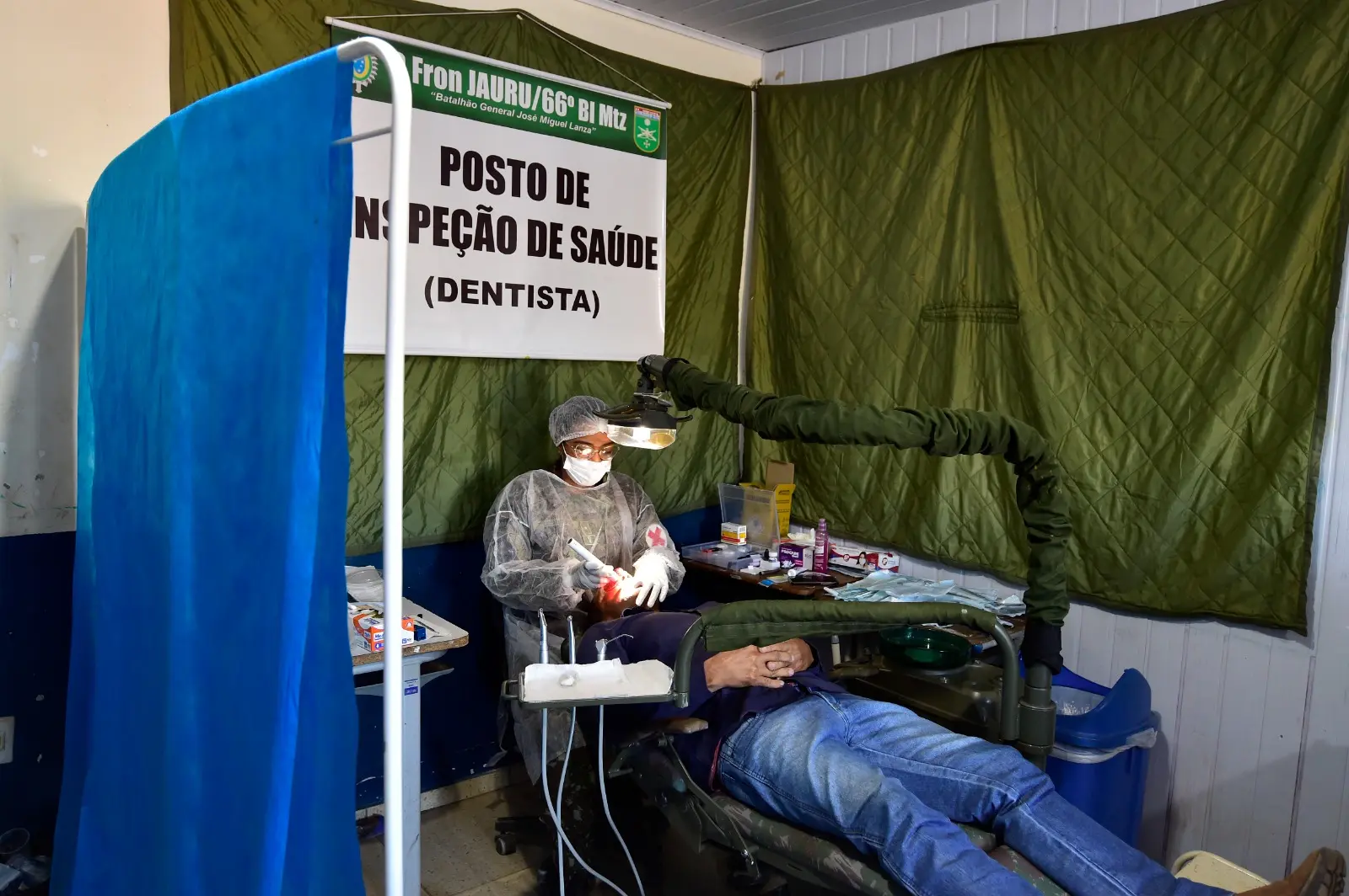 Foto que mostra a dentista do Exército, devidamente paramentada, realizando procedimento na boca de um paciente, que está deitado na cadeira odontológica.