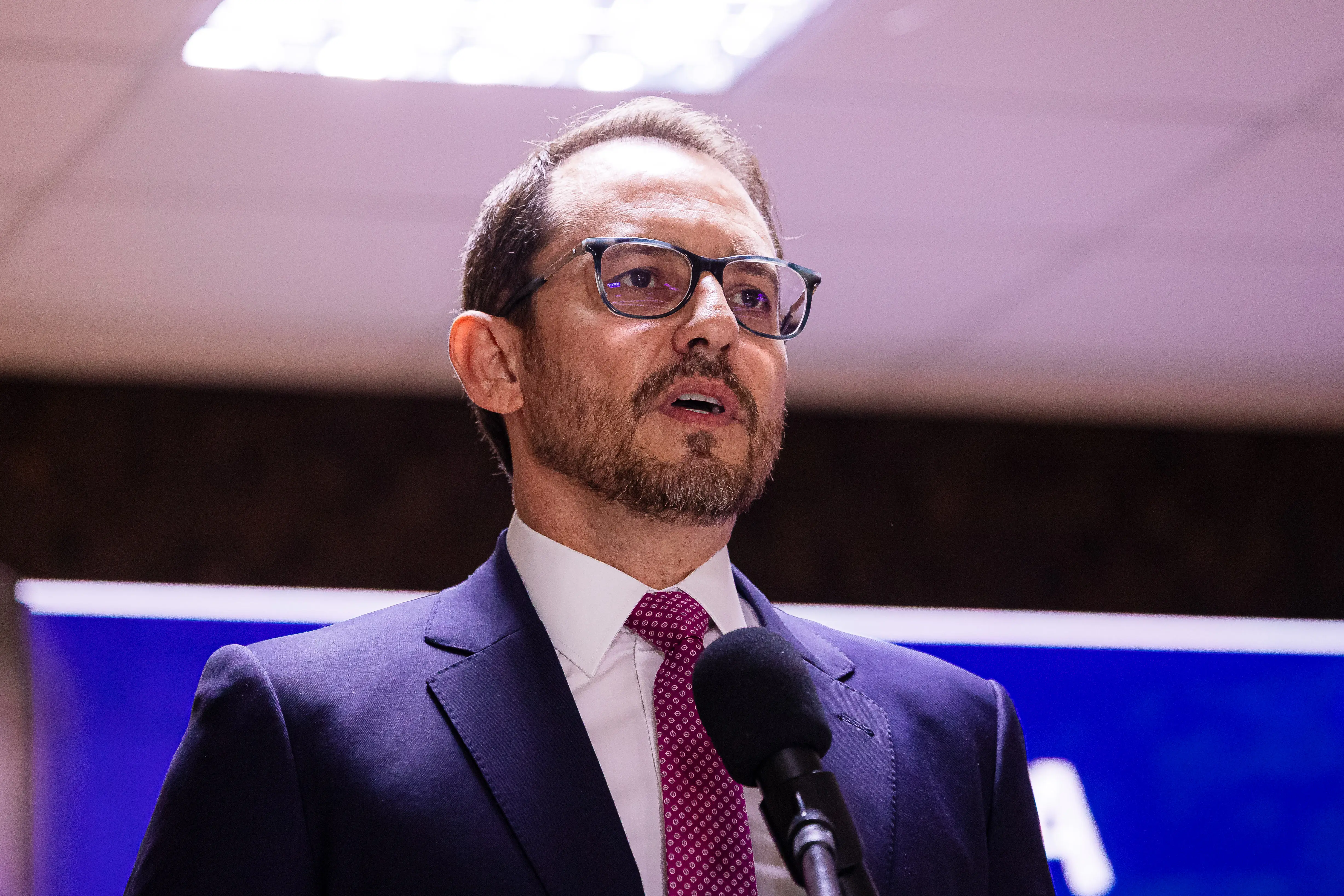 Homem branco de terno escuro, camisa branca e gravata vinho fala ao microfone. Ele tem barba aparada, cabelos curtos e usa óculos de grau. Ao fundo, um painel azul desfocado.