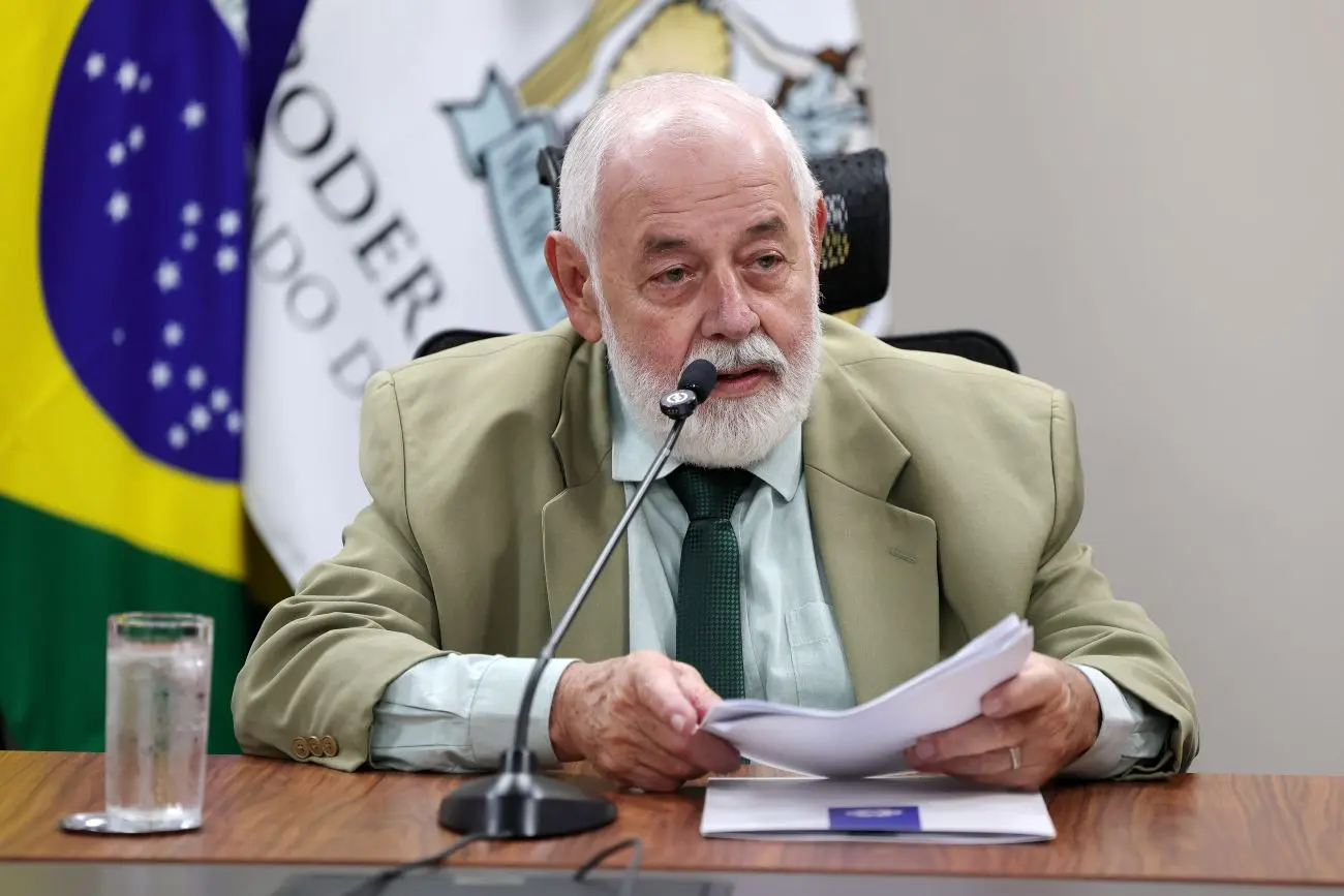 Foto horizontal que mostra o presidente do TJMT, desembargador José Zuquim Nogueira, sentado e falando ao microfone. Ele é um senhor branco, de cabelo e barba brancos, usando camisa verde clara, terno bege e gravata verde. 