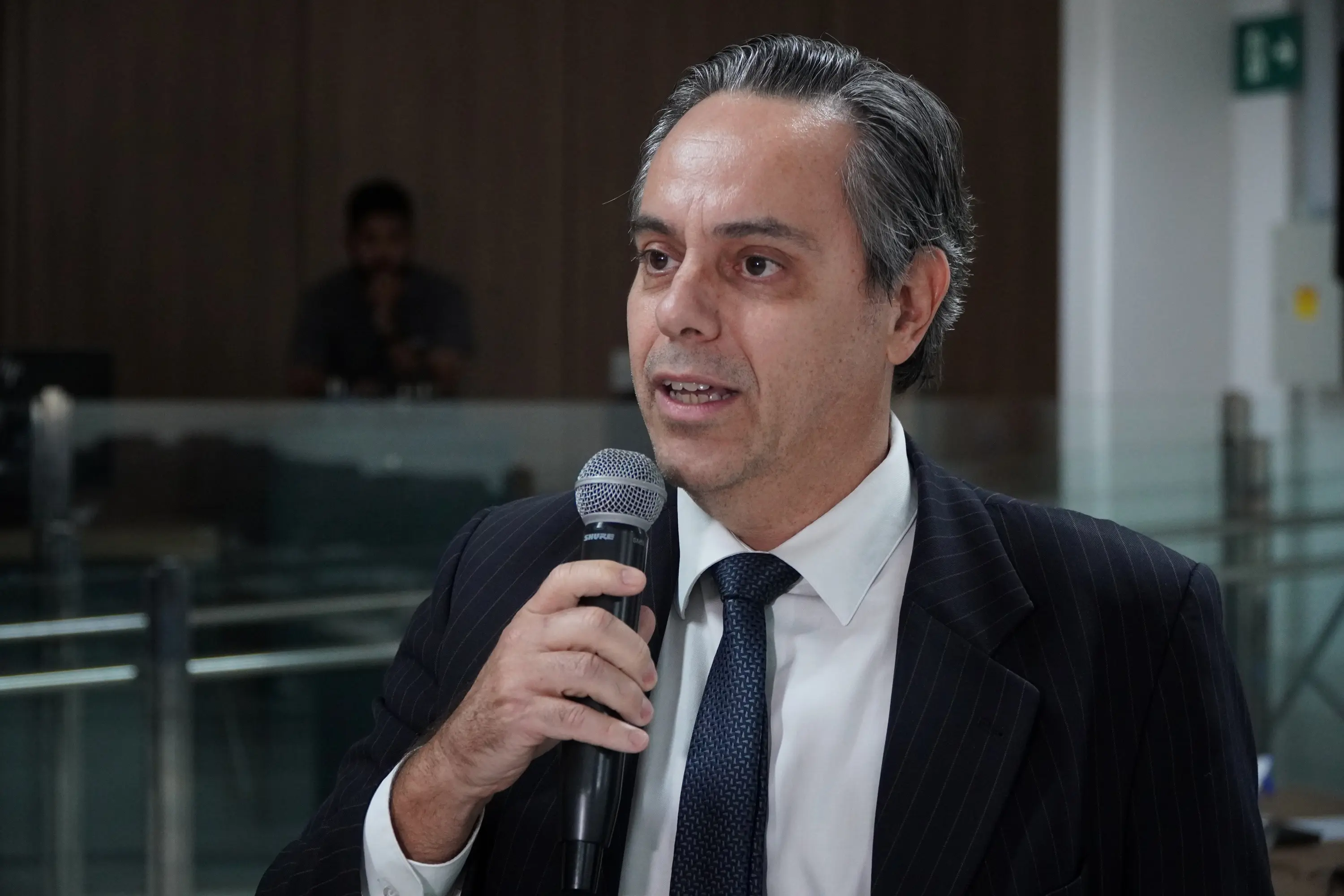 Foto horizontal que mostra o juiz Gabriel Matos (um homem de cabelos grisalhos, terno riscado escuro e gravata azul-marinho) falando ao microfone com expressão séria. Ao fundo, ambiente interno do Tribunal do Juri, com vidros e iluminação artificial.