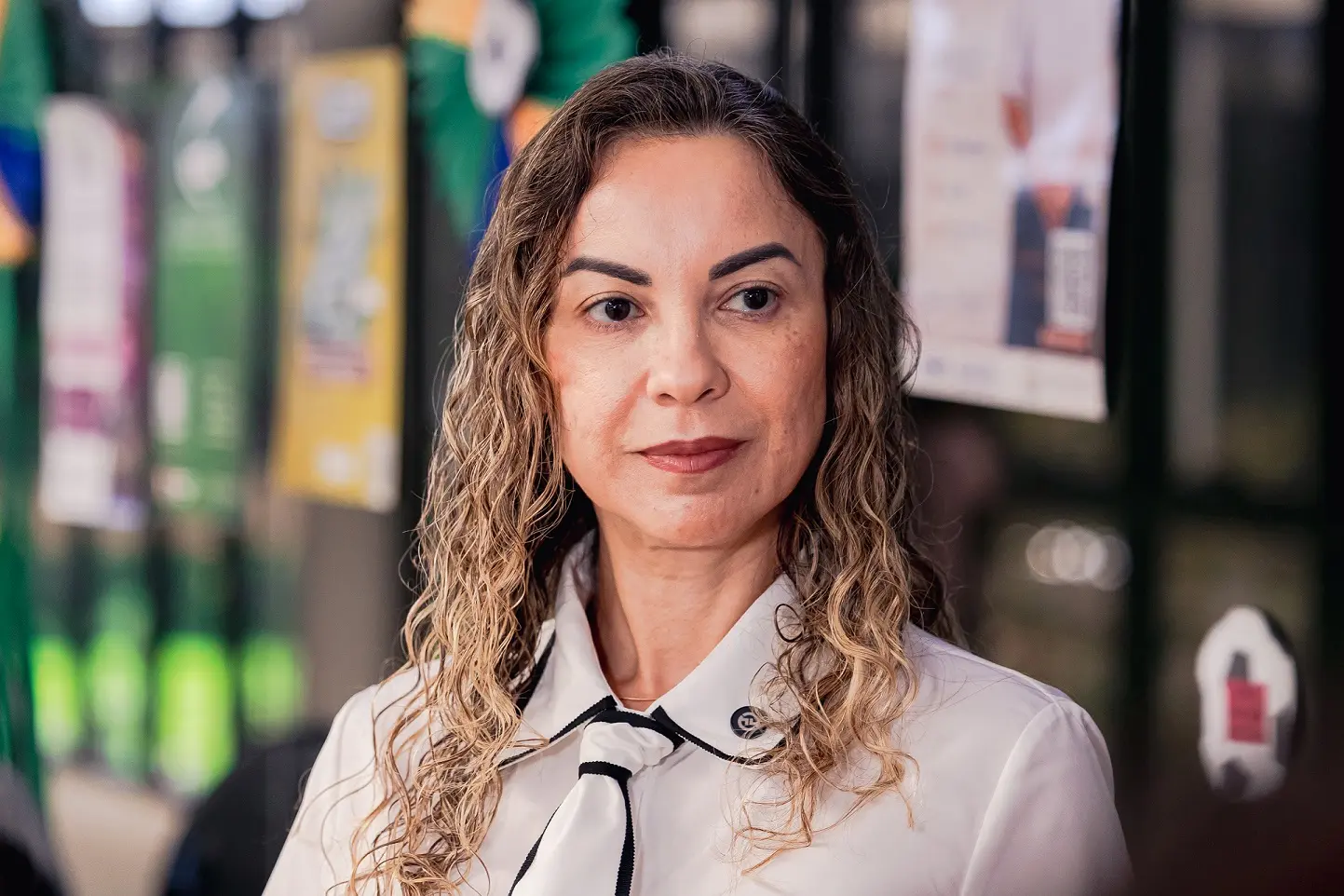 A juíza Cristiane Padim da Silva posa para fotografia em ambiente interno. A magistrada veste camisa branca com detalhes em preto e tem os cabelos cacheados na altura dos ombros. Ao fundo, aparecem painéis decorativos e elementos nas cores verde e amarelo relacionados à temática da Copa do Judiciári