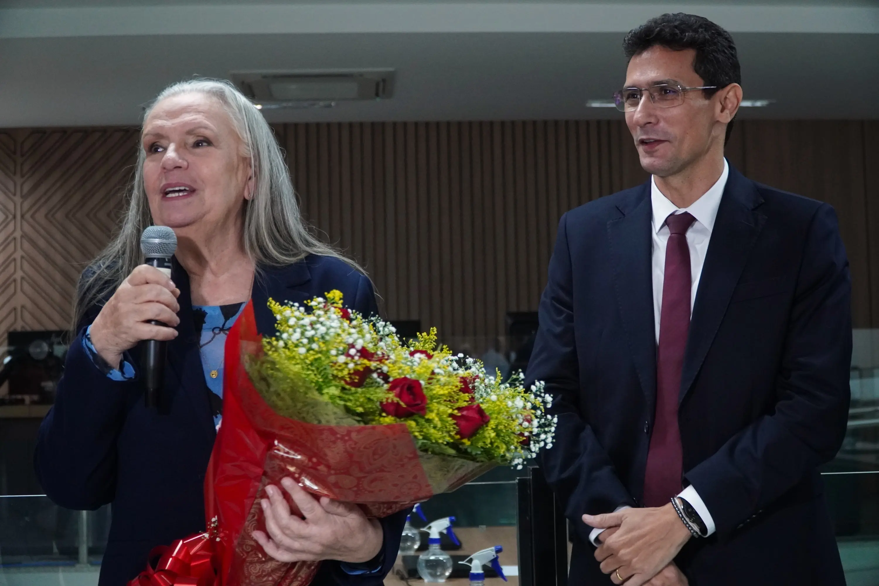 Foto horizontal que mostra a desembargadora Maria Erotides (uma mulher de cabelos longos grisalhos) segurando um buquê de rosas vermelhas e flores amarelas e falando ao microfone. Ao lado, o juiz Hugo Freitas (um homem de terno azul e gravata vinho) a observa.