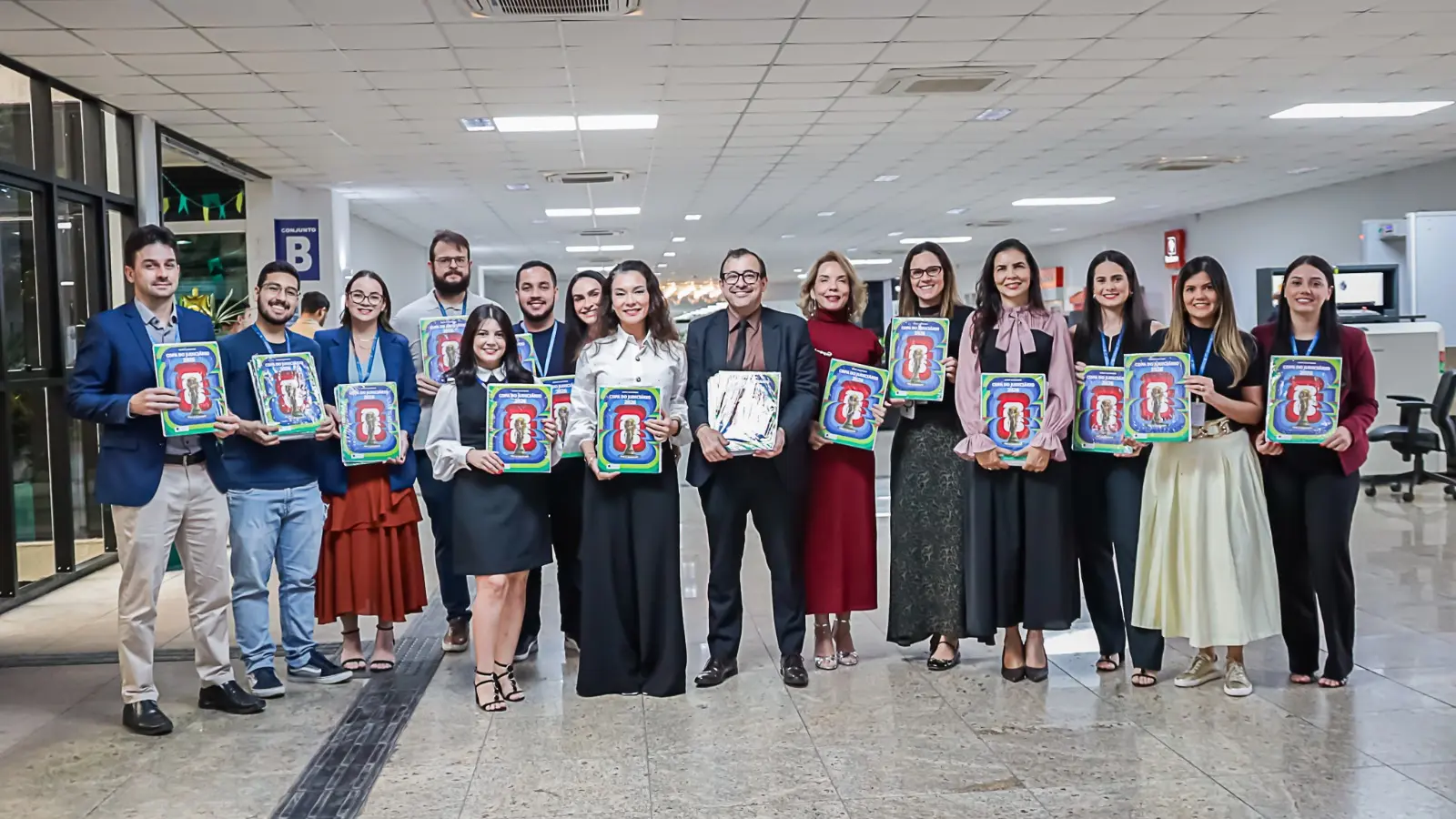 Ao centro da imagem está o juiz auxiliar da Corregedoria, Jorge Alexandre Martins Ferreira, a sua esquerda está a diretora do Fórum de Cuiabá, Hanae Yamamura de Oliveira, servidores do Fórum de Cuiabá e integrantes da equipe da Corregedoria estão de pé e posam para foto. Todos seguram um exemplar do