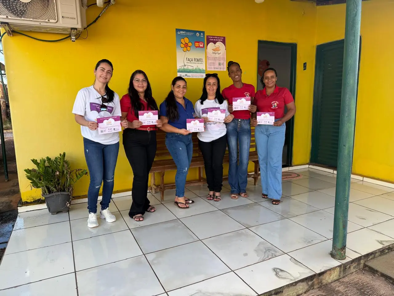 Equipe da Vara Única de Rio Branco pousa com integrantes da unidade de atendimento segurando materiais informativos da campanha. Ao fundo, na parede amarela do local, estão afixados cartazes institucionais, entre eles um com a mensagem 