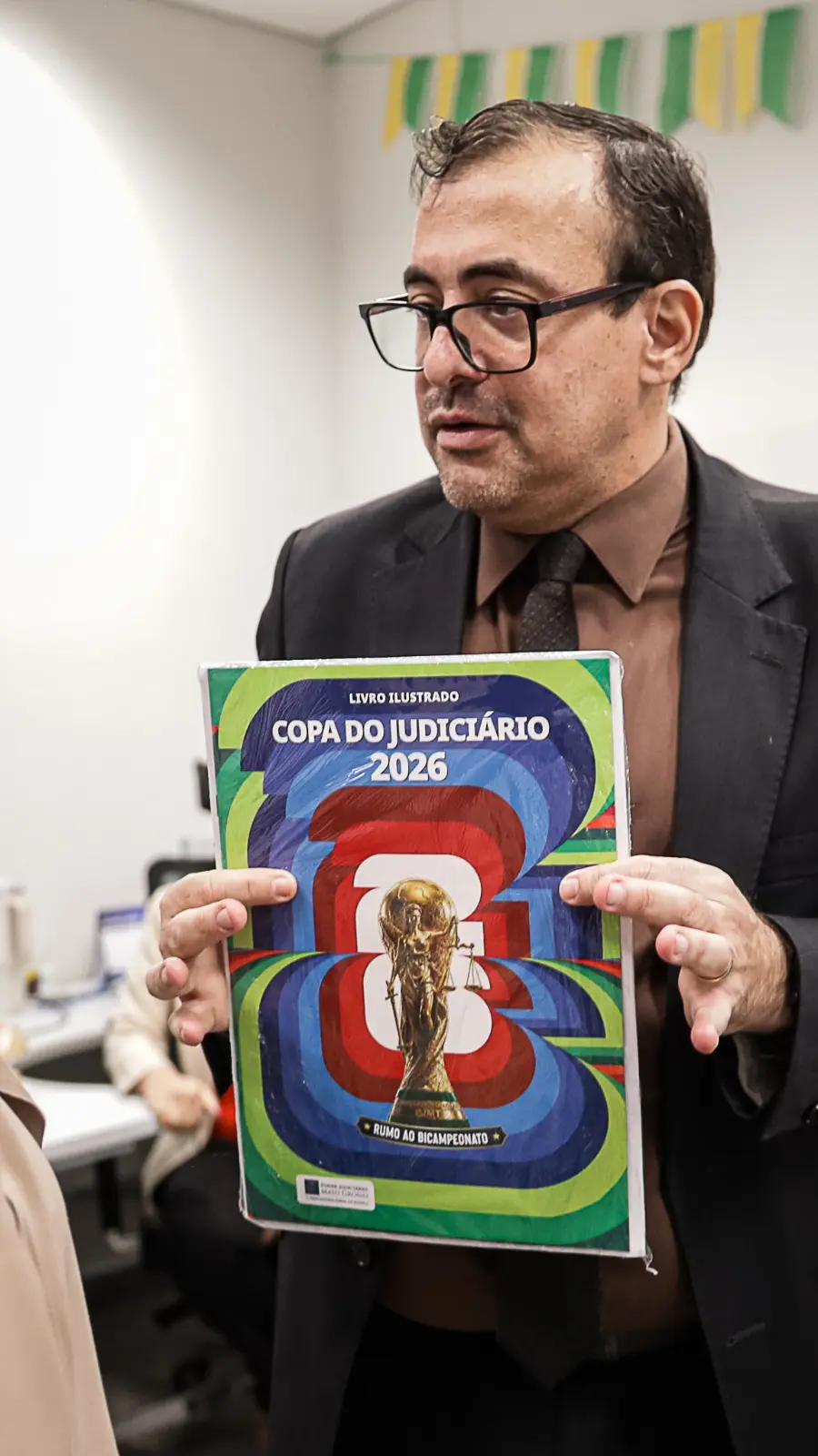 O juiz auxiliar, Jorge Alexandre Martins Ferreira, segura um exemplar do álbum físico da Copa do Judiciário 2026 durante visita a uma unidade judiciária. O magistrado veste terno escuro, camisa marrom e gravata, enquanto apresenta o material aos participantes. Ao fundo, há decoração nas cores verde 