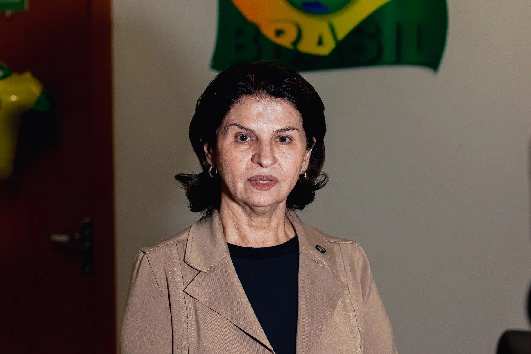 A juíza Valdeci Moraes Siqueira posa para fotografia em ambiente interno. A magistrada veste blazer bege sobre blusa preta e está posicionada em frente a uma parede decorada com elementos nas cores da bandeira do Brasil, relacionados à temática da Copa do Judiciário.