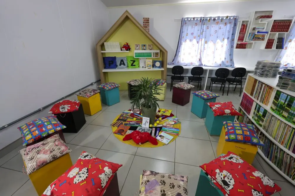 Foto horizontal que mostra uma sala de leitura onde é realizado o círculo de construção de paz na EMEB Aleixo Schenatto, em Sinop. A sala tem prateleiras com livros, pufs organizados em círculo e uma casinha de madeira para exposição de livros.