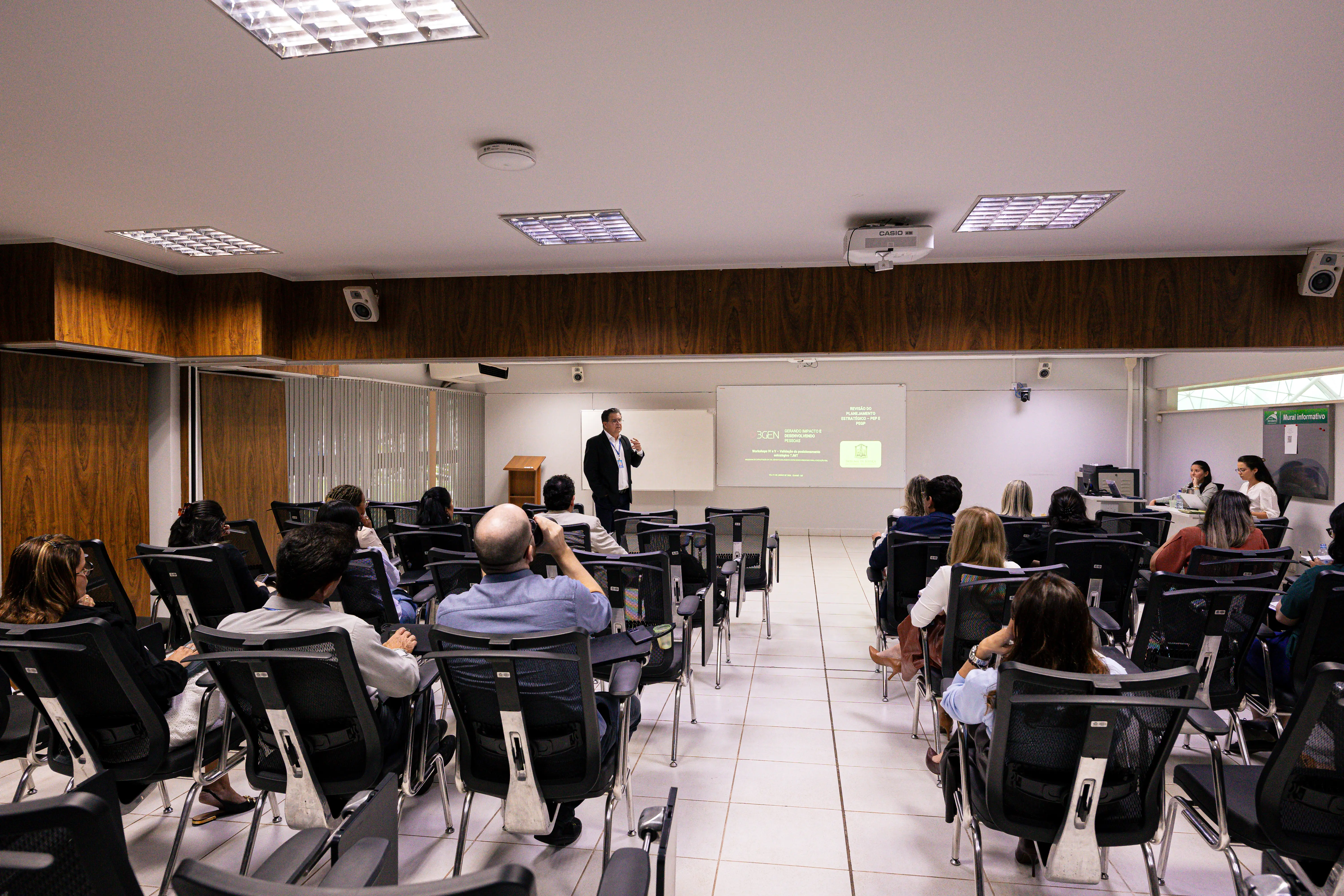 Homem em pé diante de uma tela de projeção apresenta para uma plateia sentada em cadeiras pretas dispostas em fileiras, vista de trás. Sala de teto branco, piso claro e detalhes em madeira.