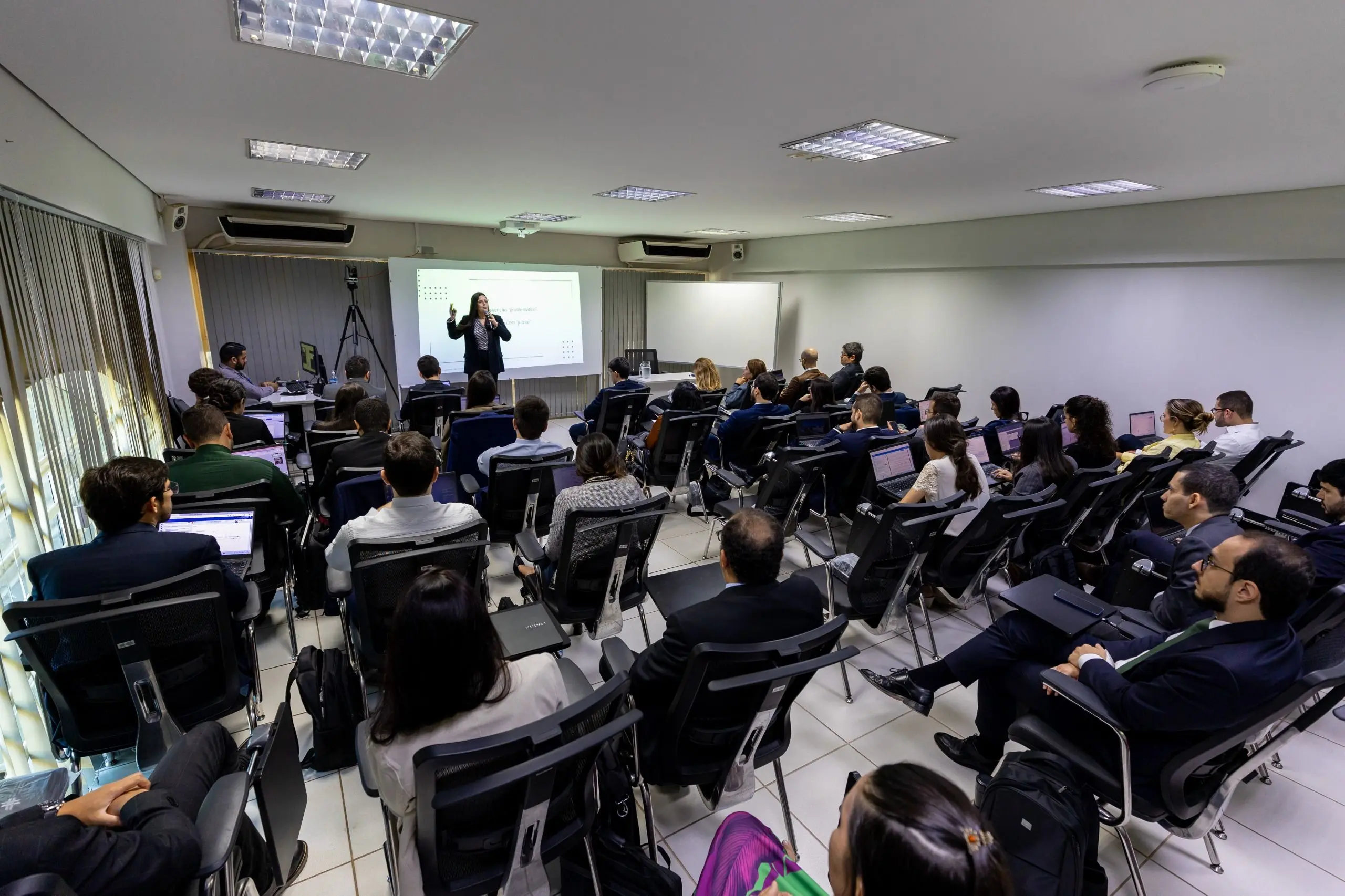 Vista do fundo de uma sala de aula cheia de alunos sentados com notebooks. À frente, uma mulher em pé fala ao lado de uma tela de projeção branca com slides em um ambiente com luz artificial.