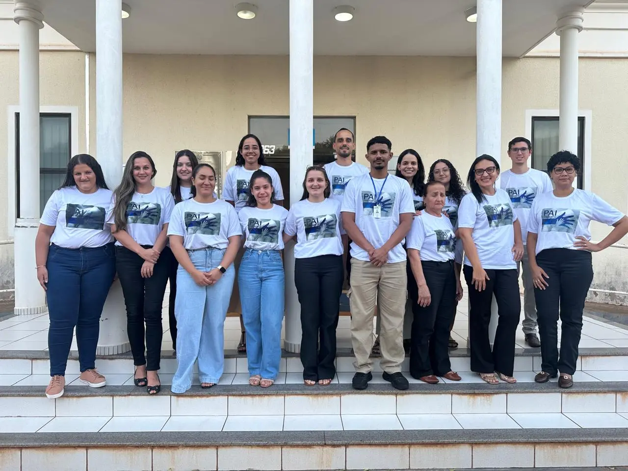 Grupo de pessoas sorri alinhado nas escadas da entrada de um prédio. Todas vestem camisetas brancas padronizadas com a estampa da campanha 