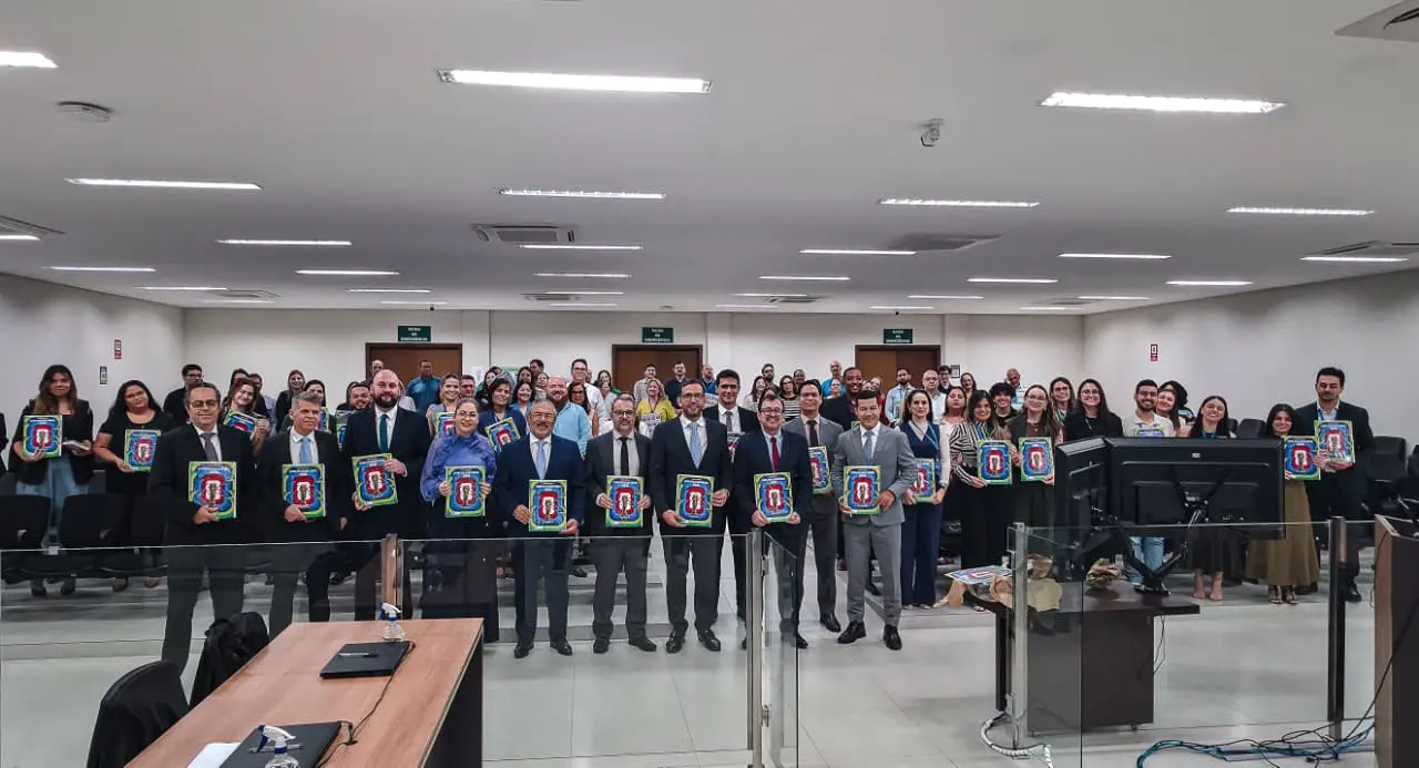 Magistrados, servidores do Fórum de Várzea Grande e a equipe da Corregedoria posam para uma fotografia após a entrega dos álbuns físicos da Copa do Judiciário 2026. Na primeira fila, os magistrados seguram exemplares do álbum, enquanto servidores e demais participantes aparecem ao fundo, também exib