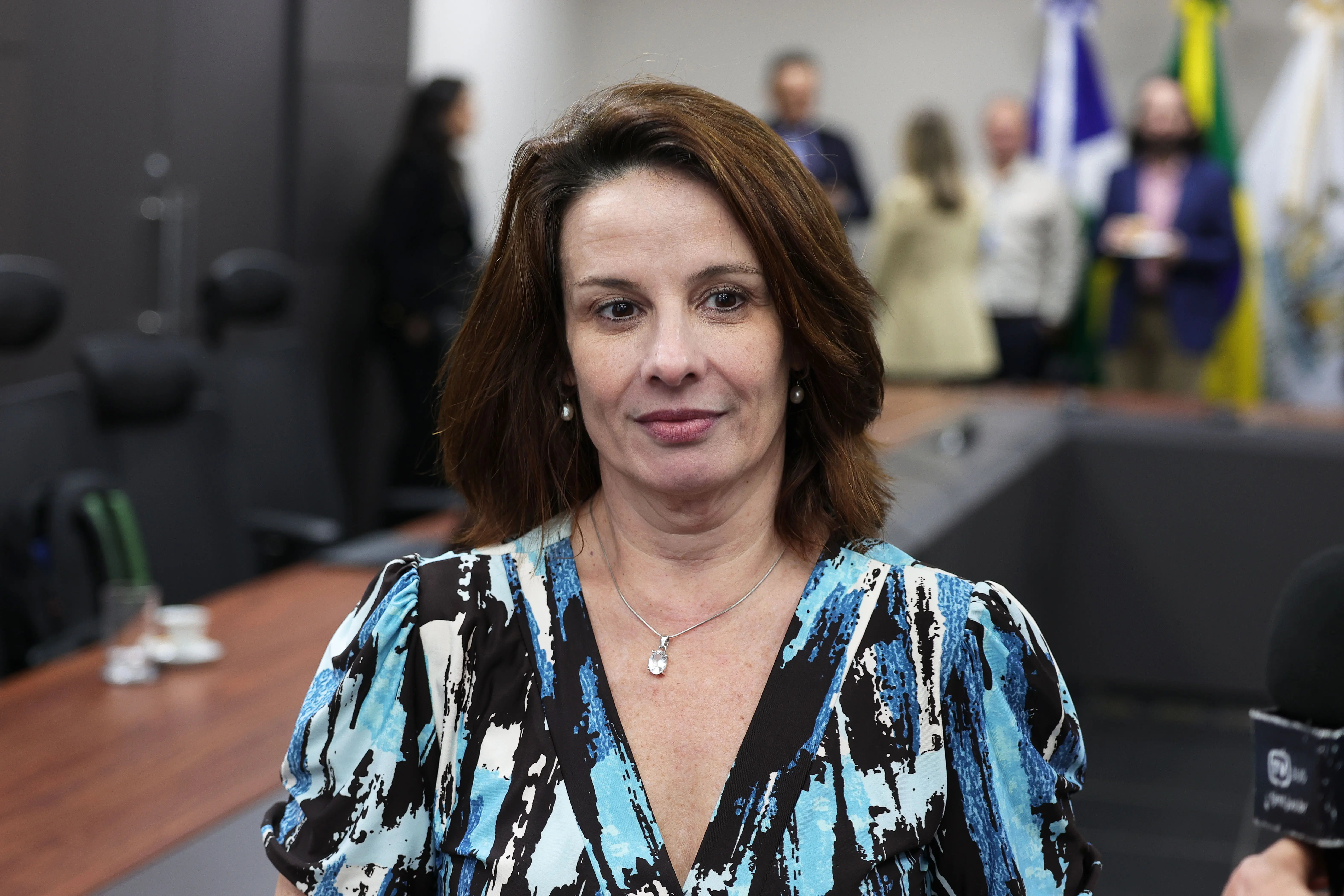 Mulher de cabelos castanhos, veste blusa estampada em azul, preto e branco, com colar de pingente, é fotografada em plano frontal. Ao fundo, pessoas em pé e bandeiras desfocadas.