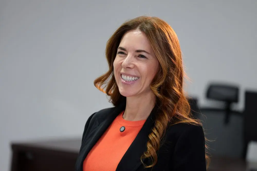 Foto horizontal que mostra a juíza Débora Caldas do busto pra cima, sorrindo. Ela é uma mulher branca, magra, de cabelos longos e castanhos, usando blusa laranjada e blazer preto.