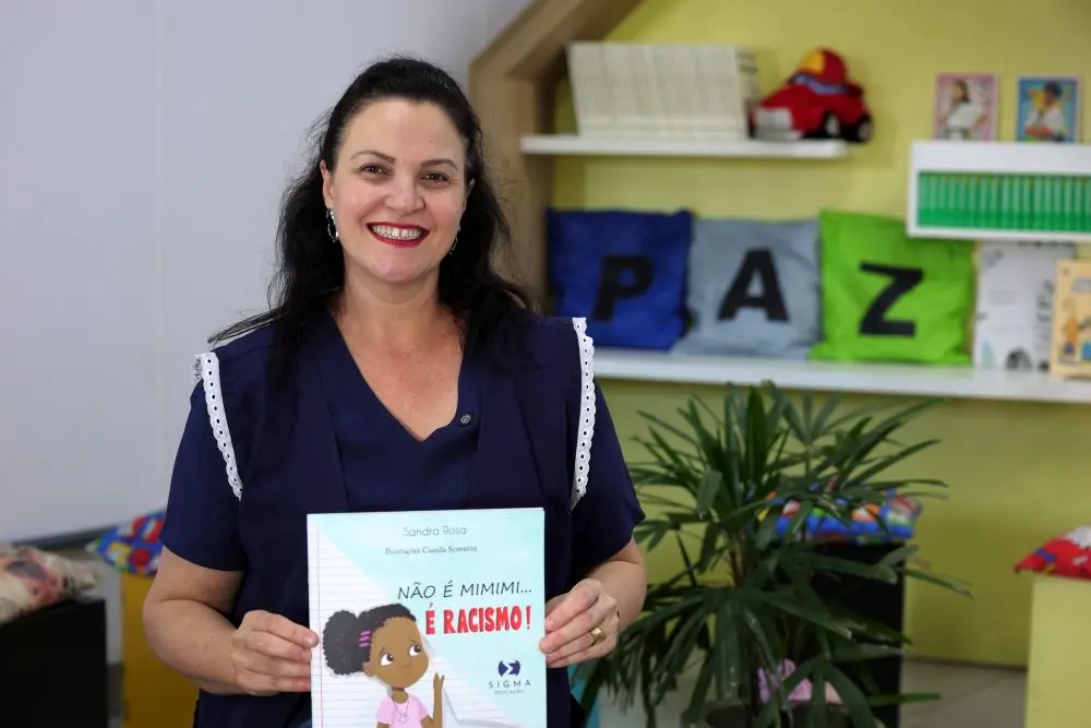 Foto horizontal que mostra a professora Djordana Cecília Bombarda , da cintura pra cima, sorrindo para a foto e mostrando a capa de um livro chamado