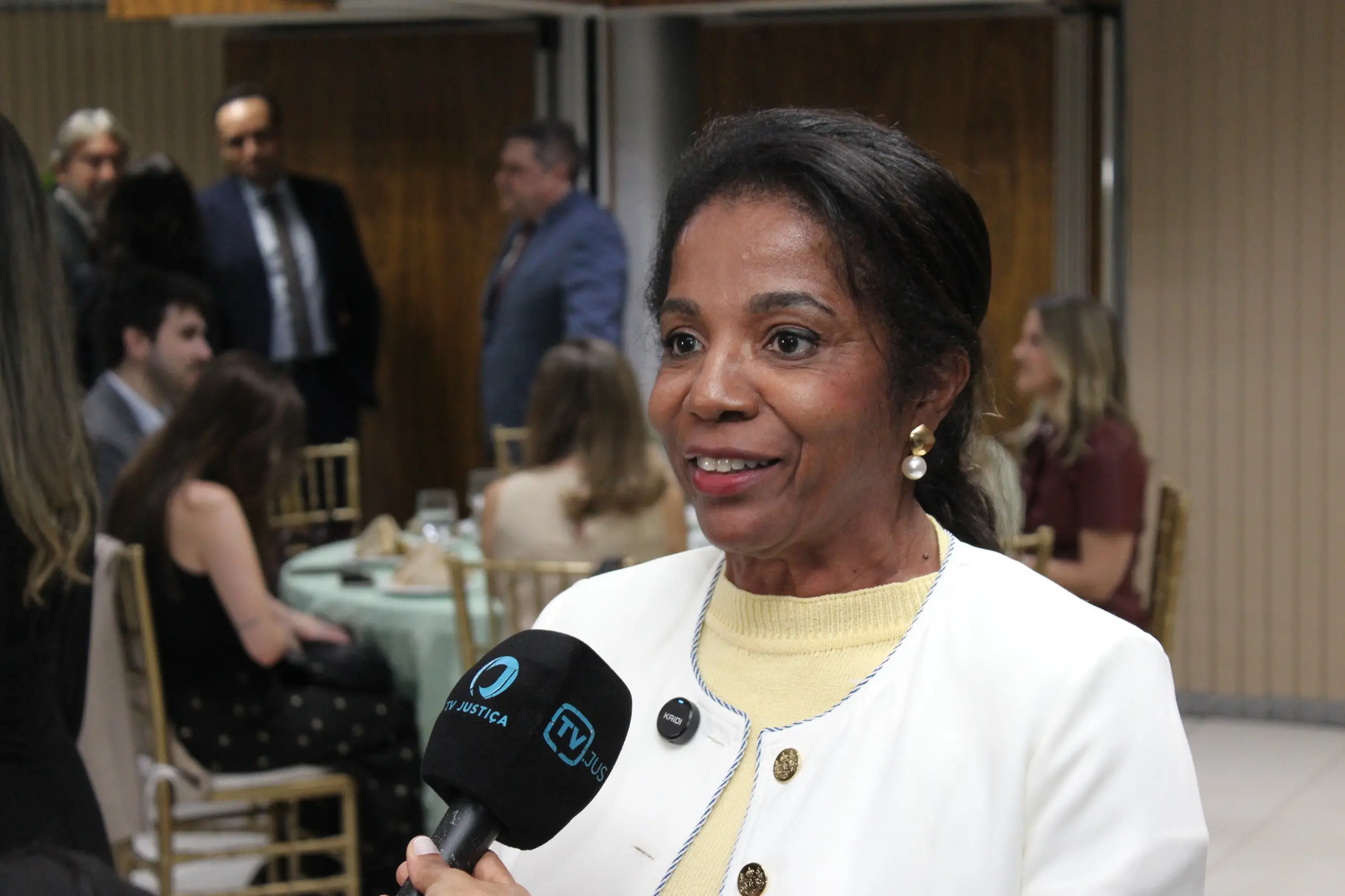 Pessoa em traje formal branco concede entrevista segurando um microfone com logotipo, em ambiente interno. Ao fundo, há mesas com pessoas sentadas, outras em pé e uma parede com revestimento de madeira, indicando evento institucional.