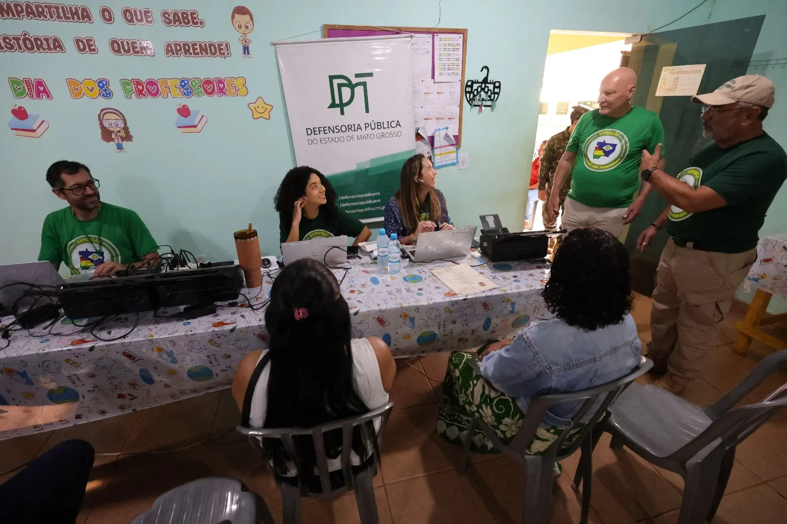 Atendimento da Defensoria Pública de MT em sala decorada. Três profissionais sentados atrás de mesa com notebooks atendem duas mulheres. Dois homens em pé conversam no lado direito.