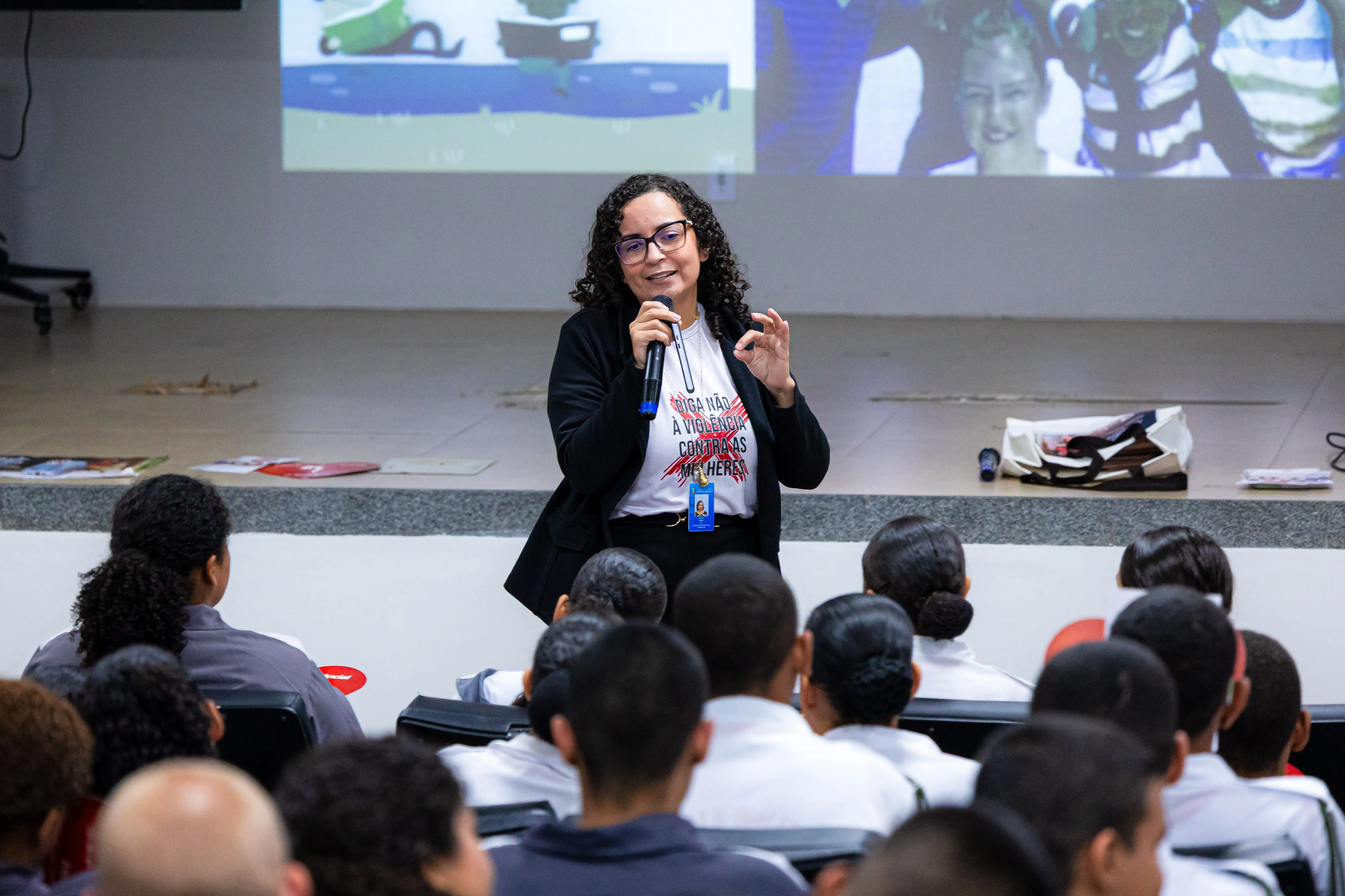 Mulher de cabelos cacheados, óculos e blazer preto sobre camiseta com a frase "Diga não à violência contra as mulheres", fala ao microfone durante palestra a estudantes.