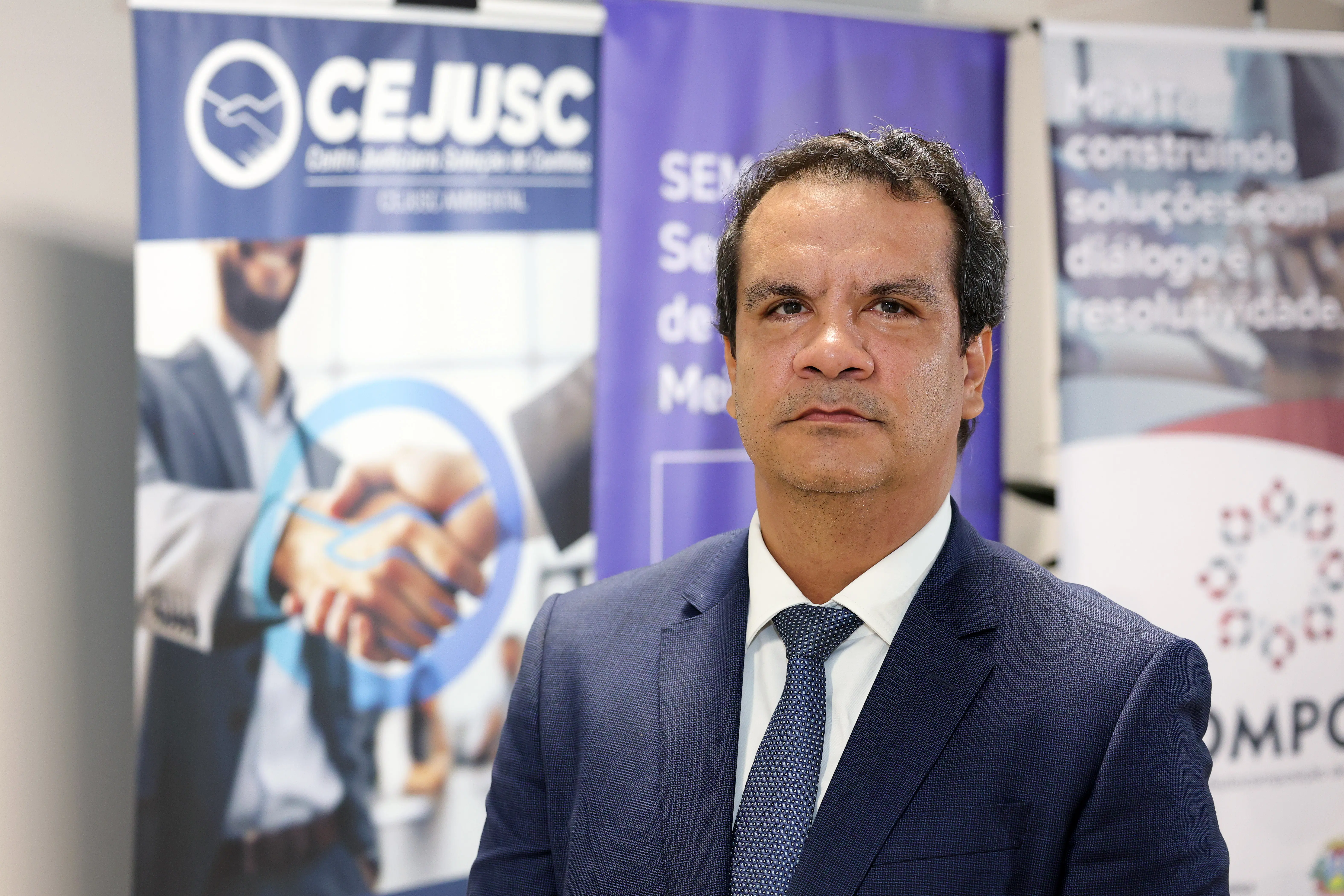 Retrato em plano médio de um homem vestindo terno azul-marinho, camisa branca e gravata texturizada azul. Ele possui expressão neutra e está posicionado à frente de banners institucionais do CEJUSC e do MPMT.