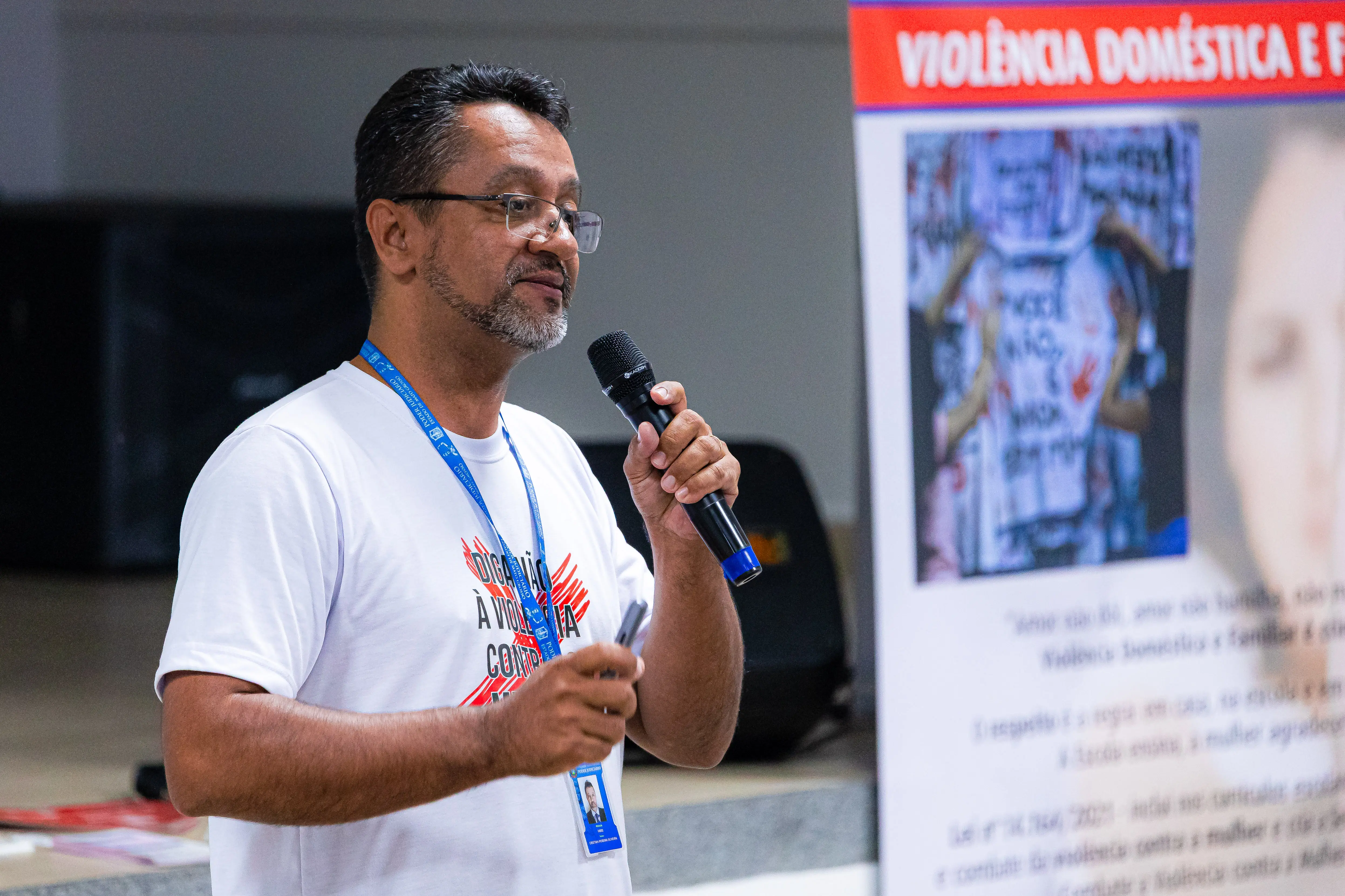 Homem de óculos, barba grisalha e camiseta branca com a frase "Diga não à violência contra as mulheres", fala ao microfone diante de banner sobre violência doméstica e familiar.